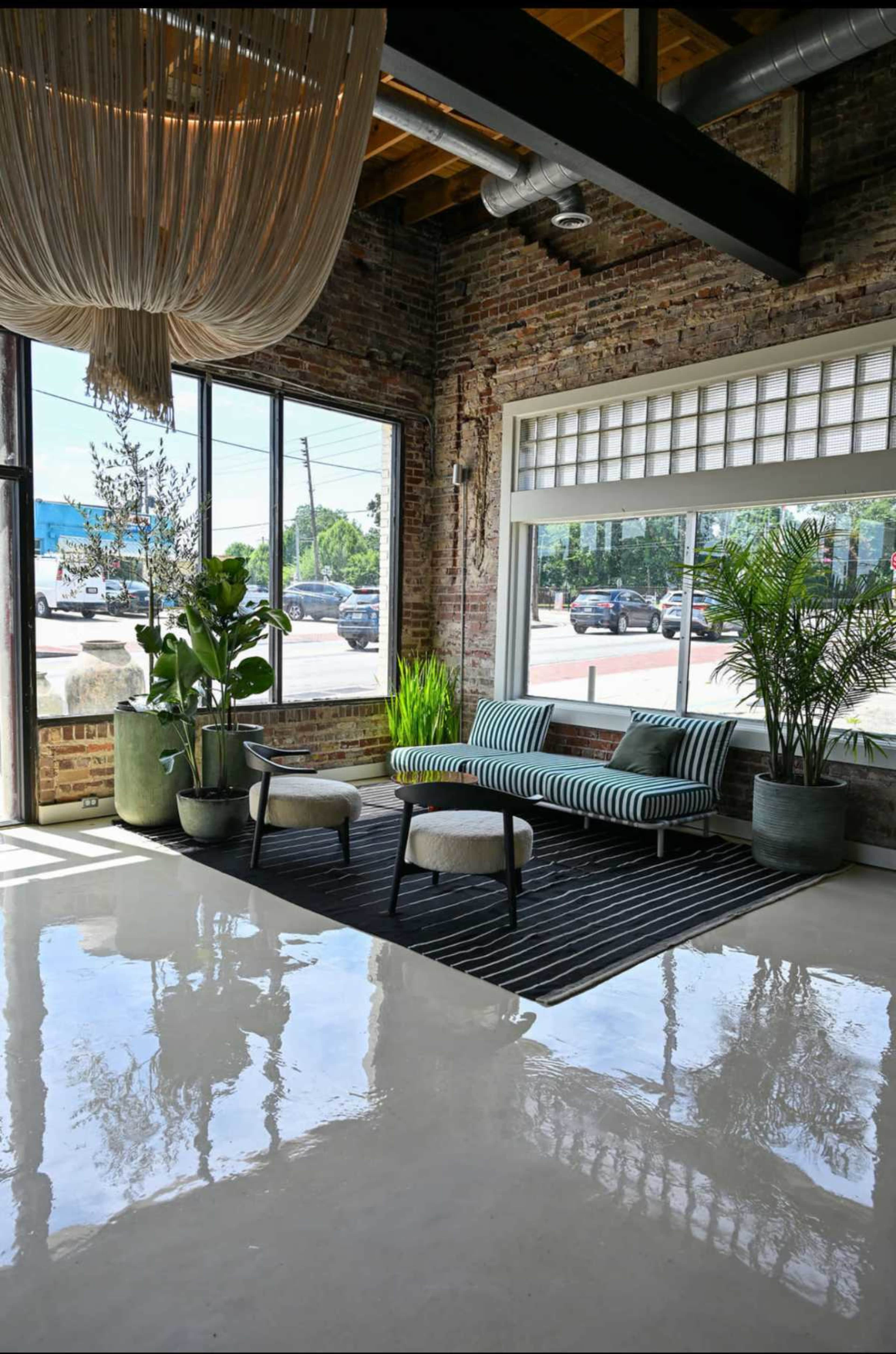 A modern lounge area features a striped sofa, two chairs, large plants, and a polished floor, with sunlight streaming through expansive windows.