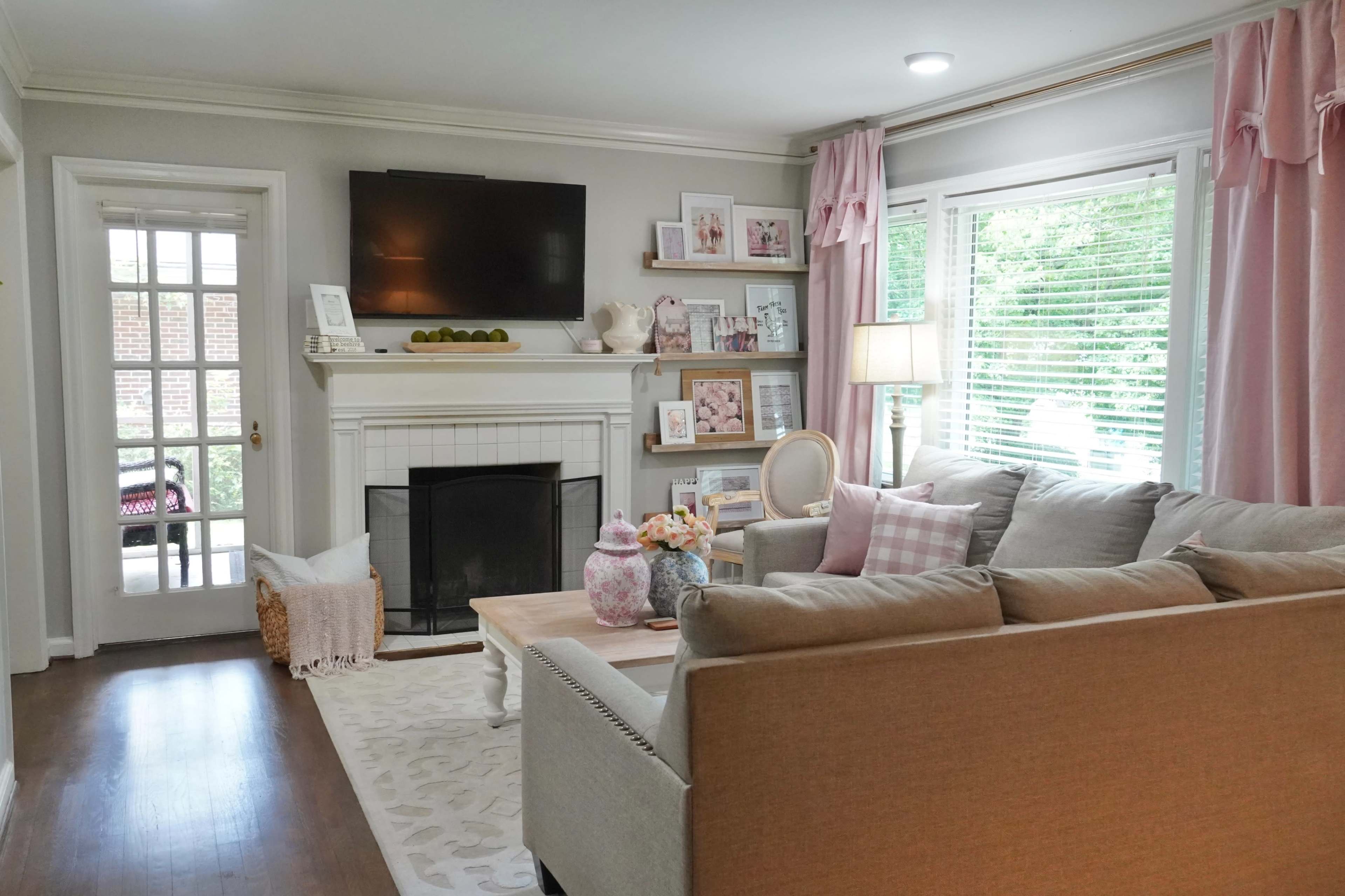 A living room with a light gray couch, wooden coffee table, and a fireplace beneath a mounted television, surrounded by windows with natural light and pink curtains.