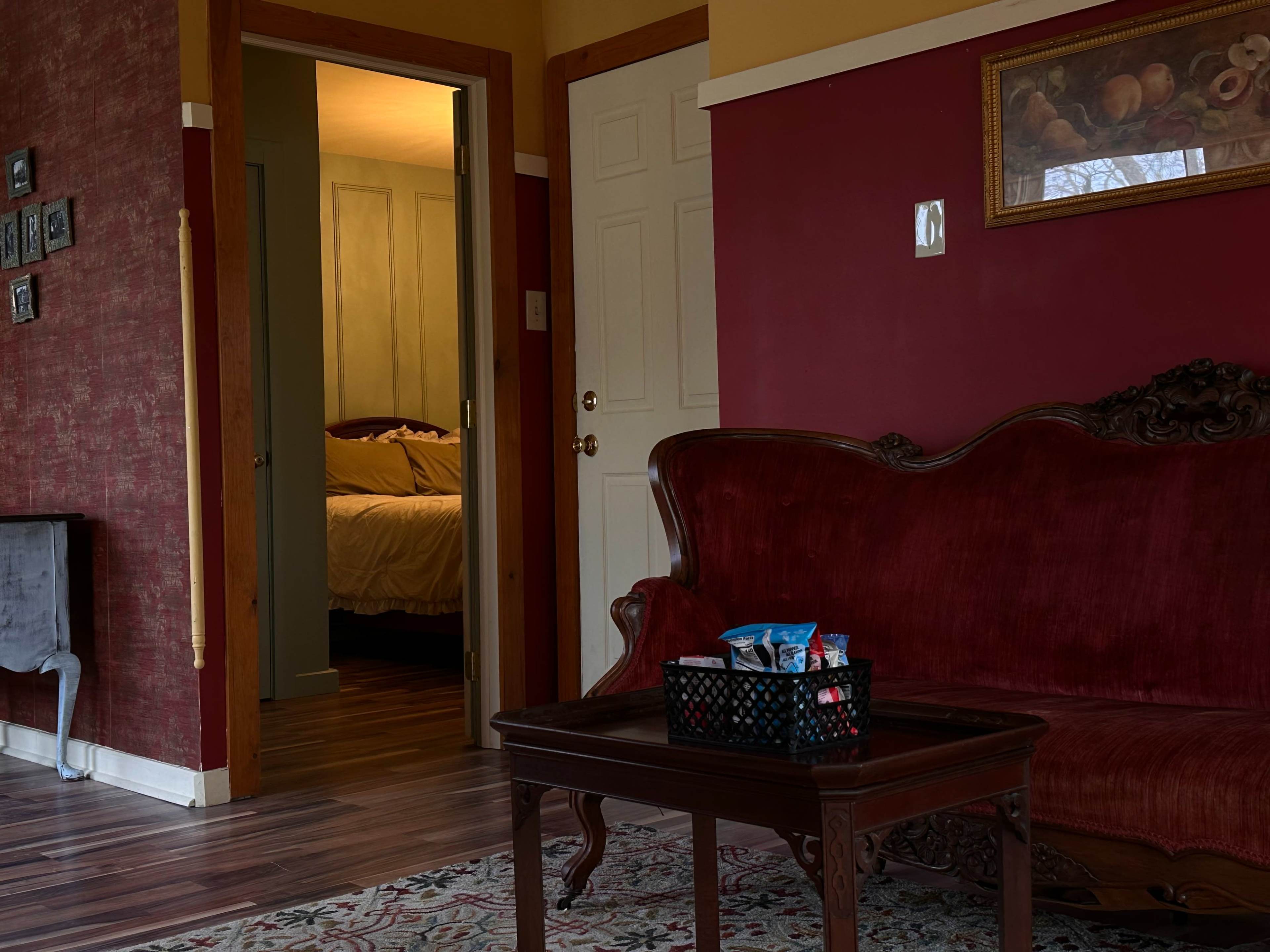 A cozy living room features a red velvet sofa and a wooden coffee table with a basket of snacks, leading to a dimly lit bedroom beyond an open door.