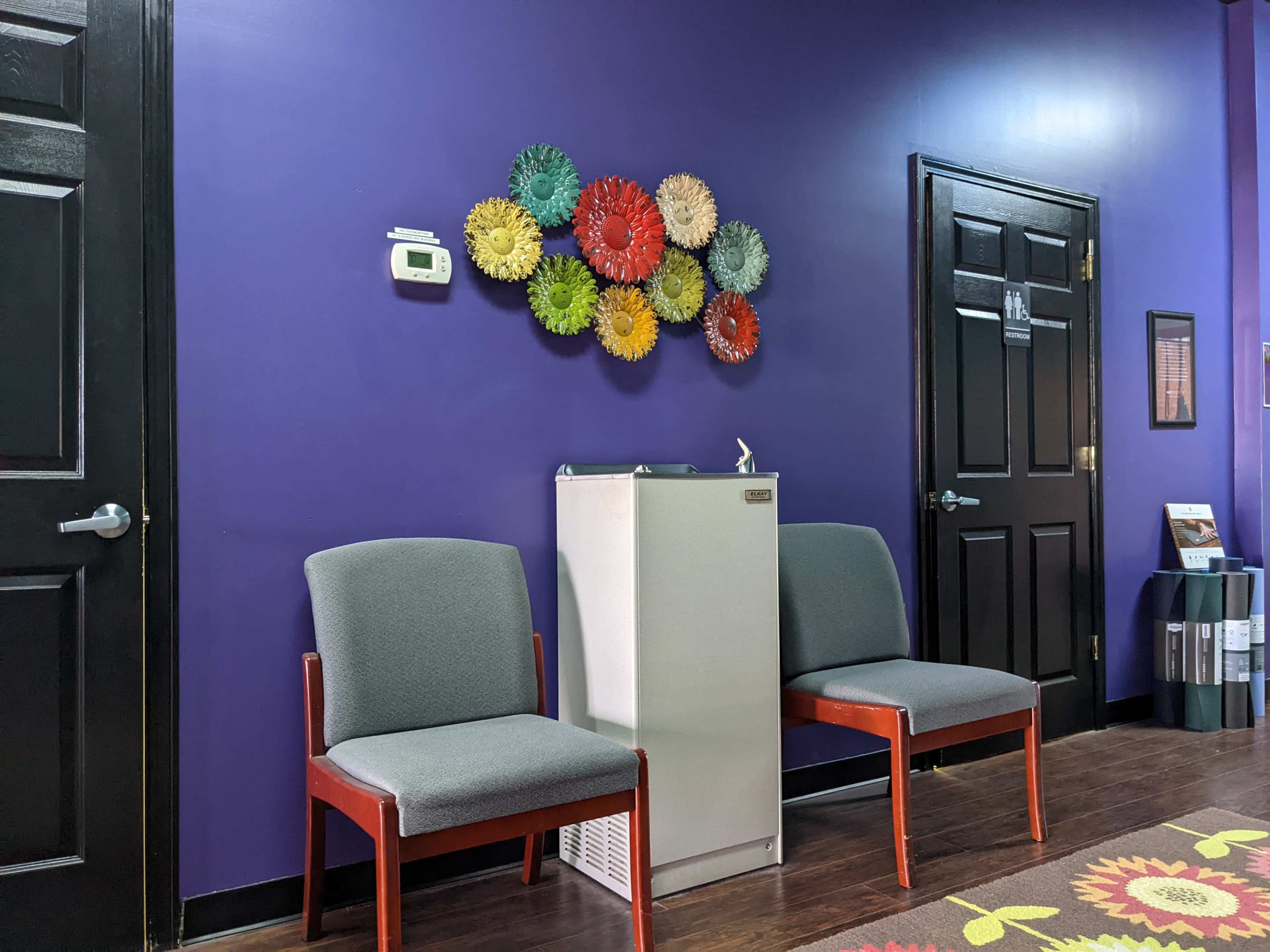 Two gray chairs are positioned next to a small fridge against a purple wall adorned with colorful circular decorations.
