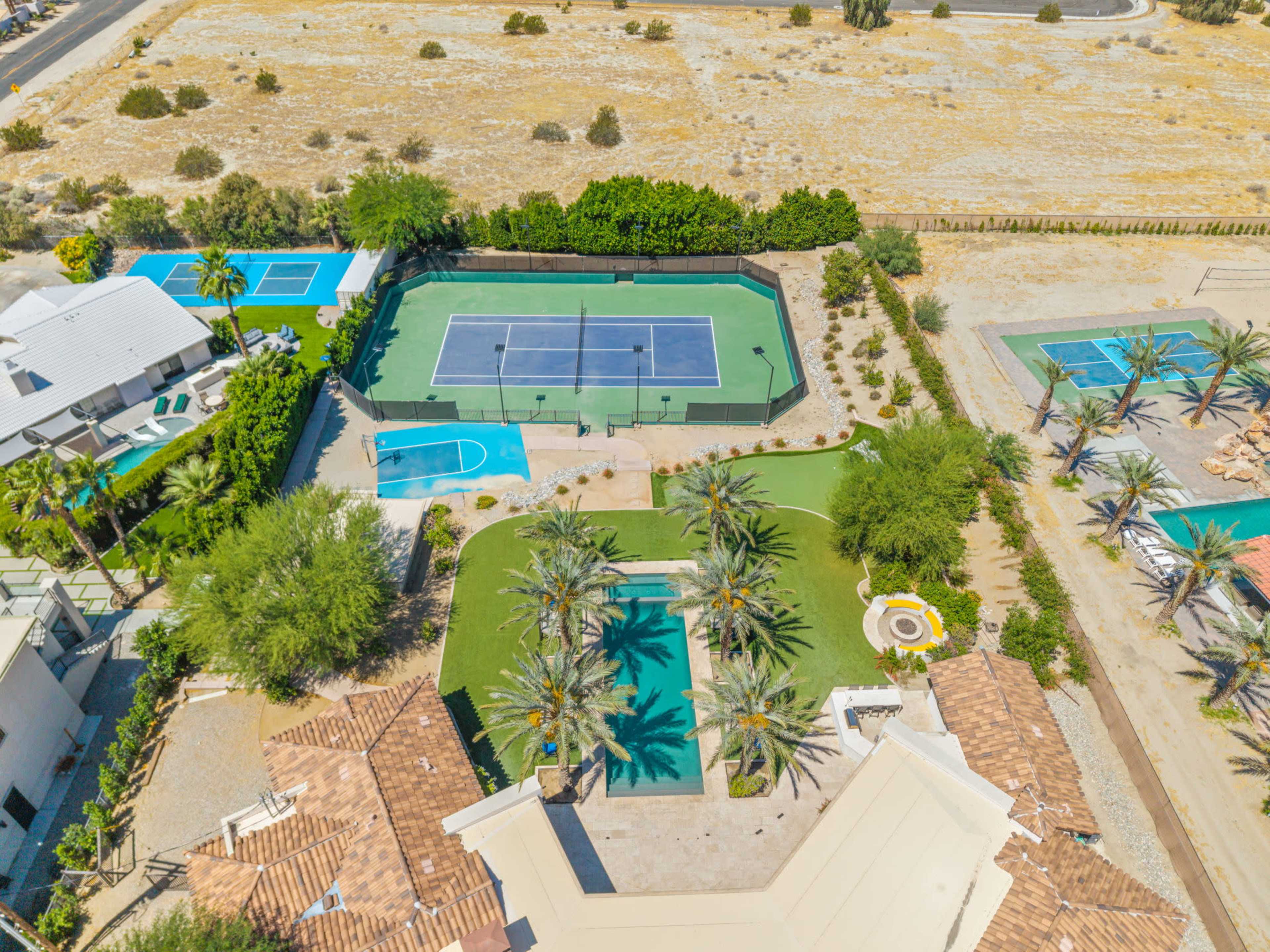The image shows an aerial view of a landscaped property featuring tennis and basketball courts, a swimming pool, and a desert landscape.