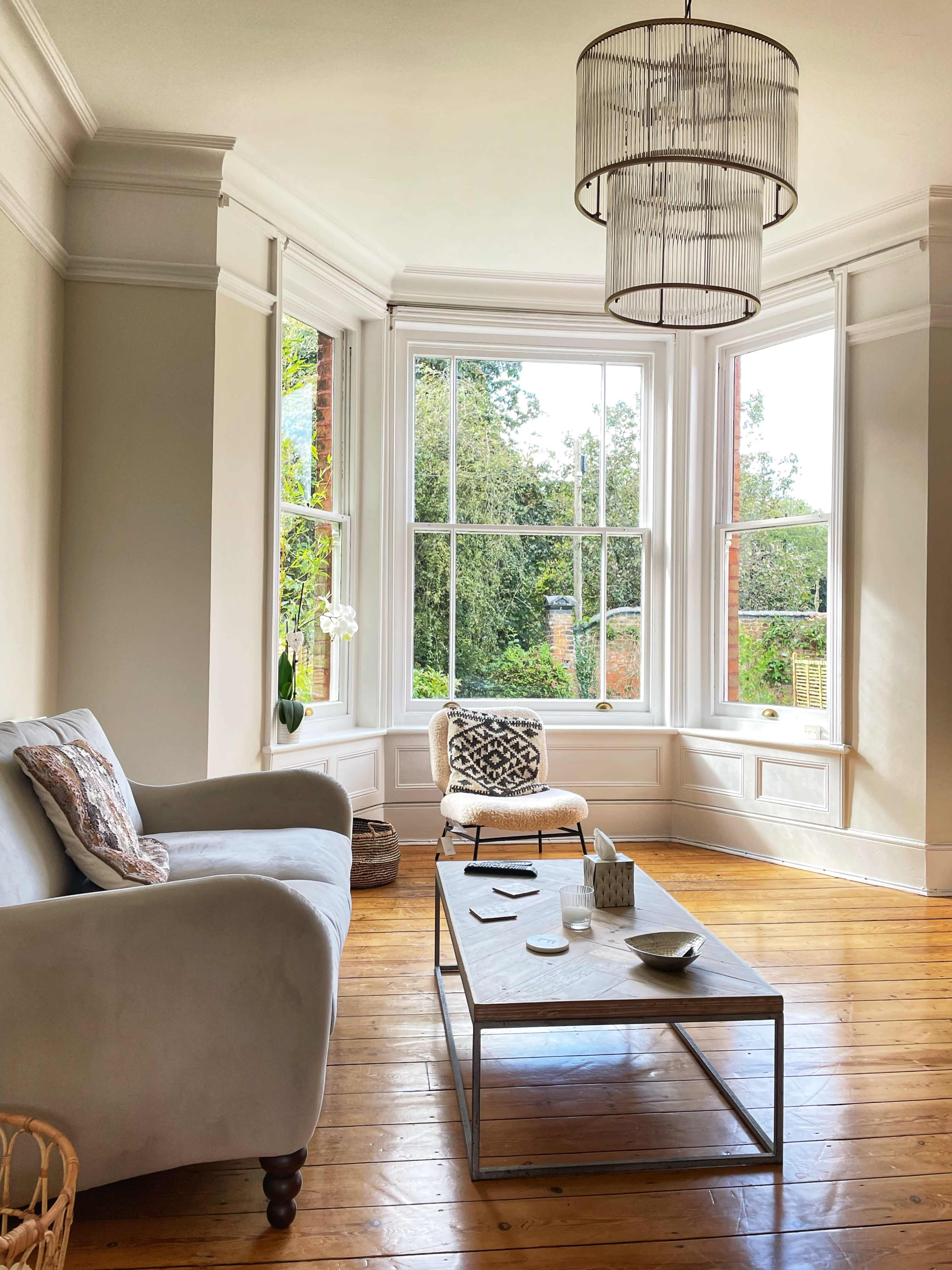 A living room features a light gray sofa, a wooden coffee table with various items, and large windows providing views of greenery outside.