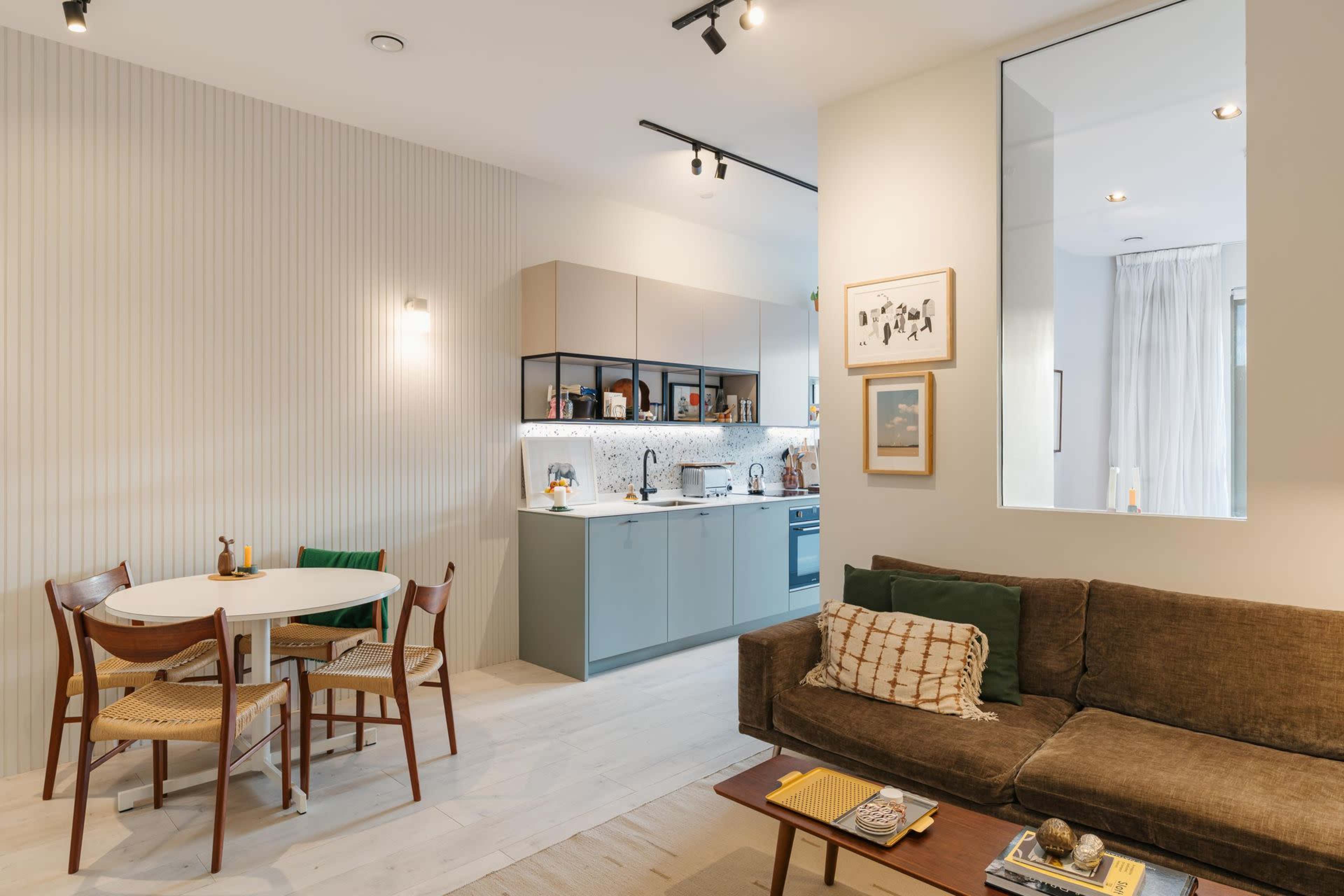The image shows a modern living space featuring a kitchen area with light cabinetry, a dining table with four chairs, and a cozy seating area with a sofa.