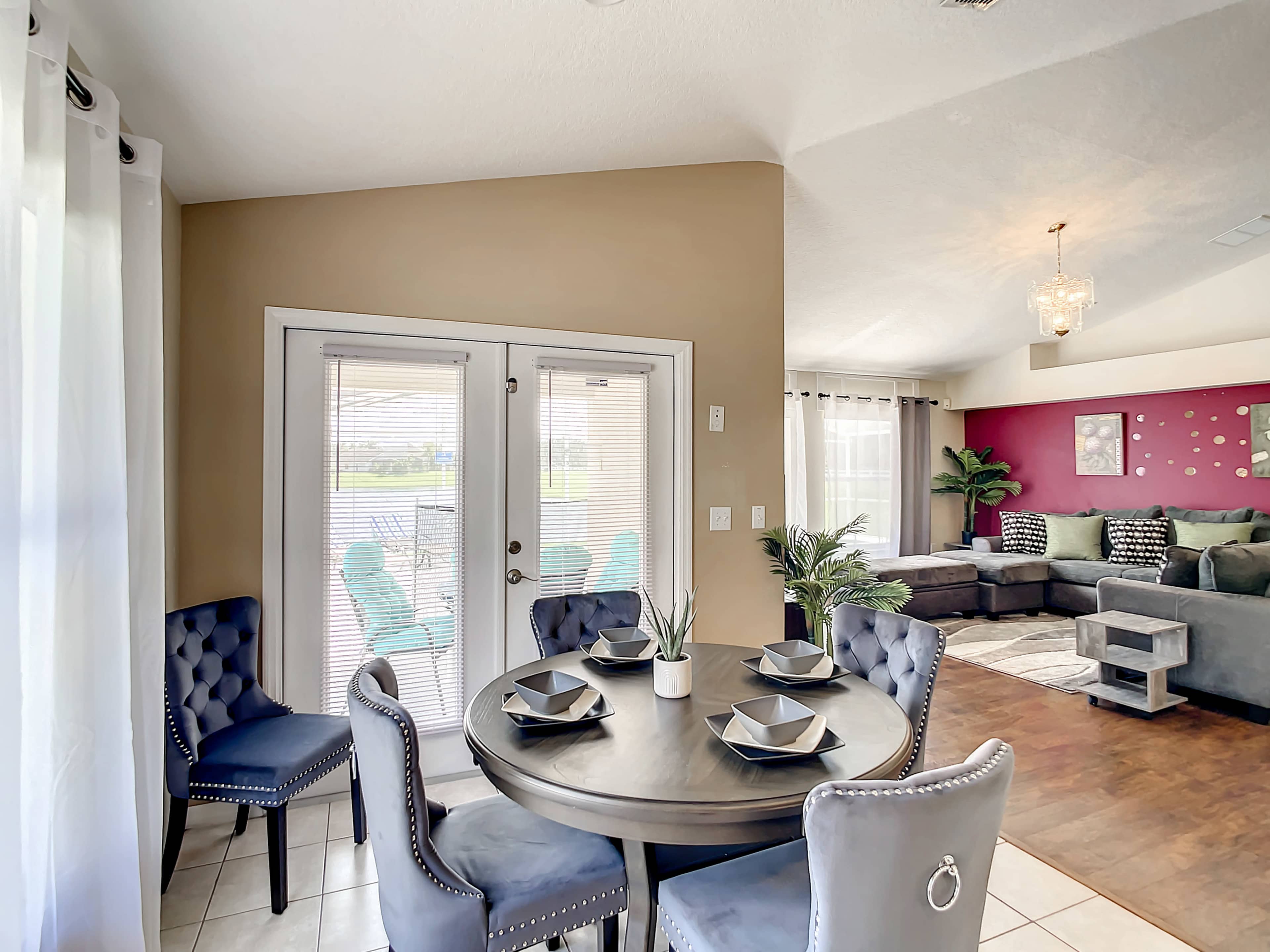 A dining area with a round table surrounded by four upholstered chairs, adjacent to a living room featuring a sectional sofa and large windows.