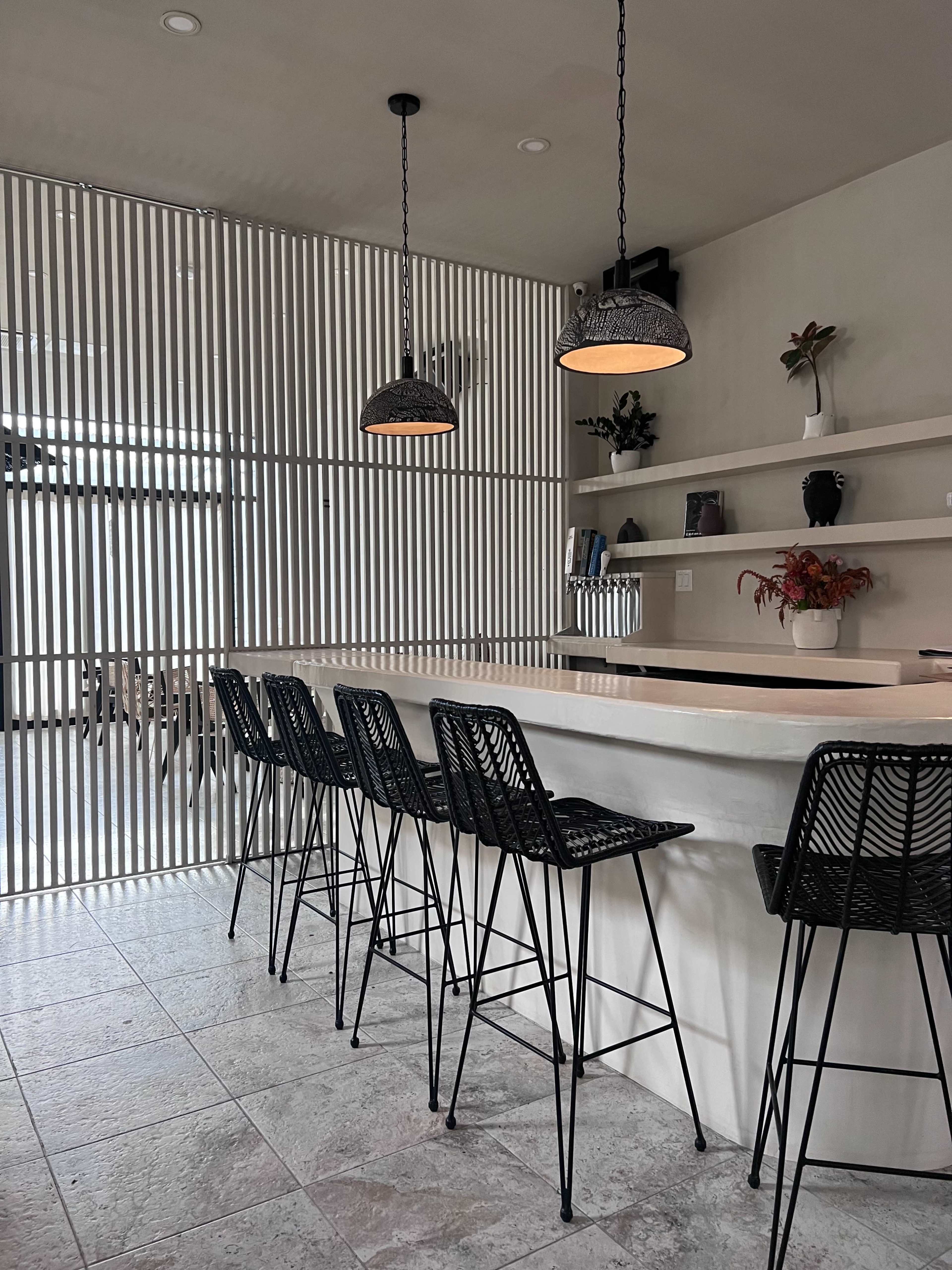 The image shows a modern kitchen bar area with a curved countertop and six black wire bar stools, illuminated by two hanging light fixtures, set against a backdrop of vertical slatted walls.