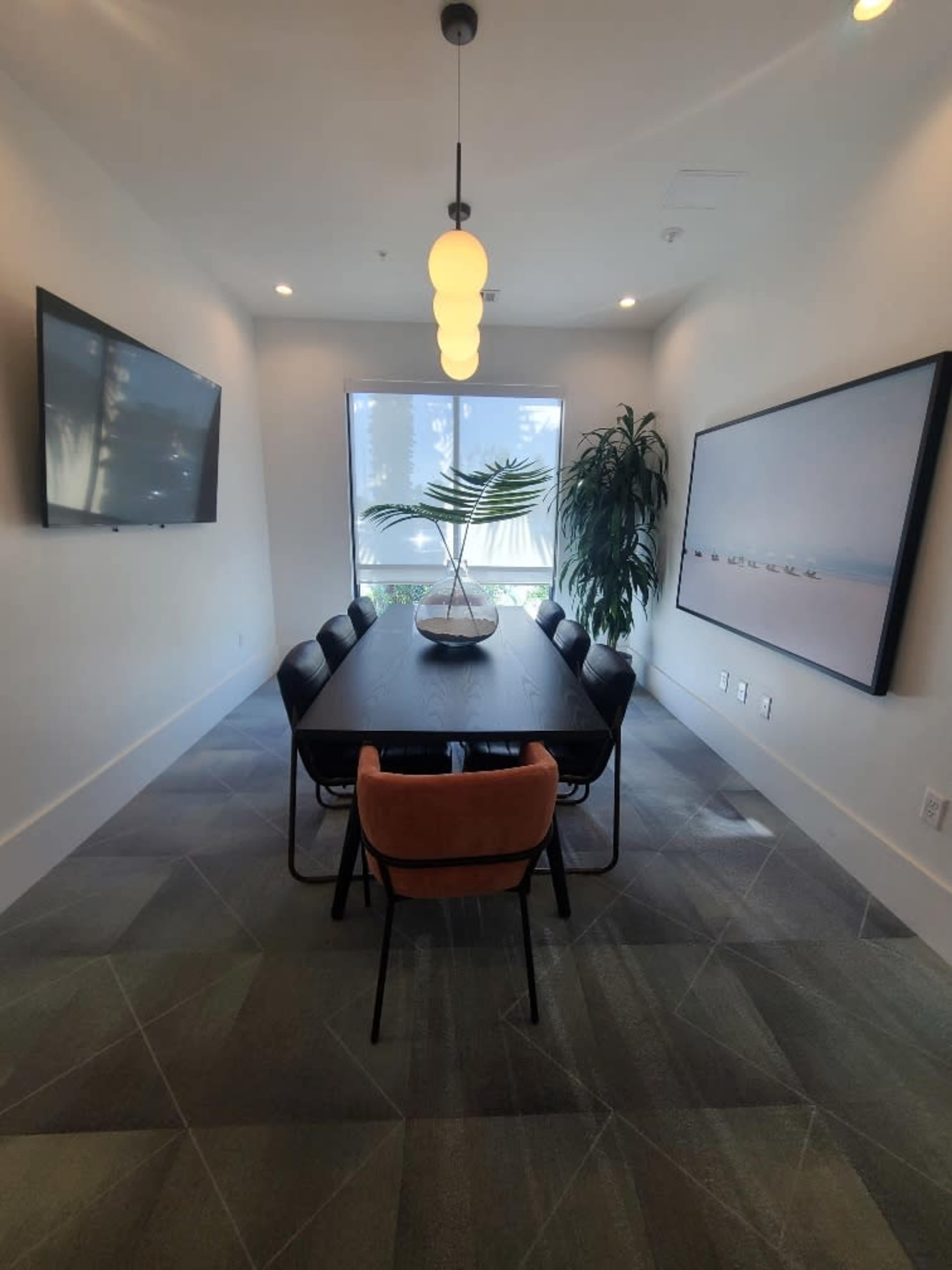 A long black table with black chairs and one orange chair is set in a bright meeting room featuring two televisions and a large window with a potted plant.