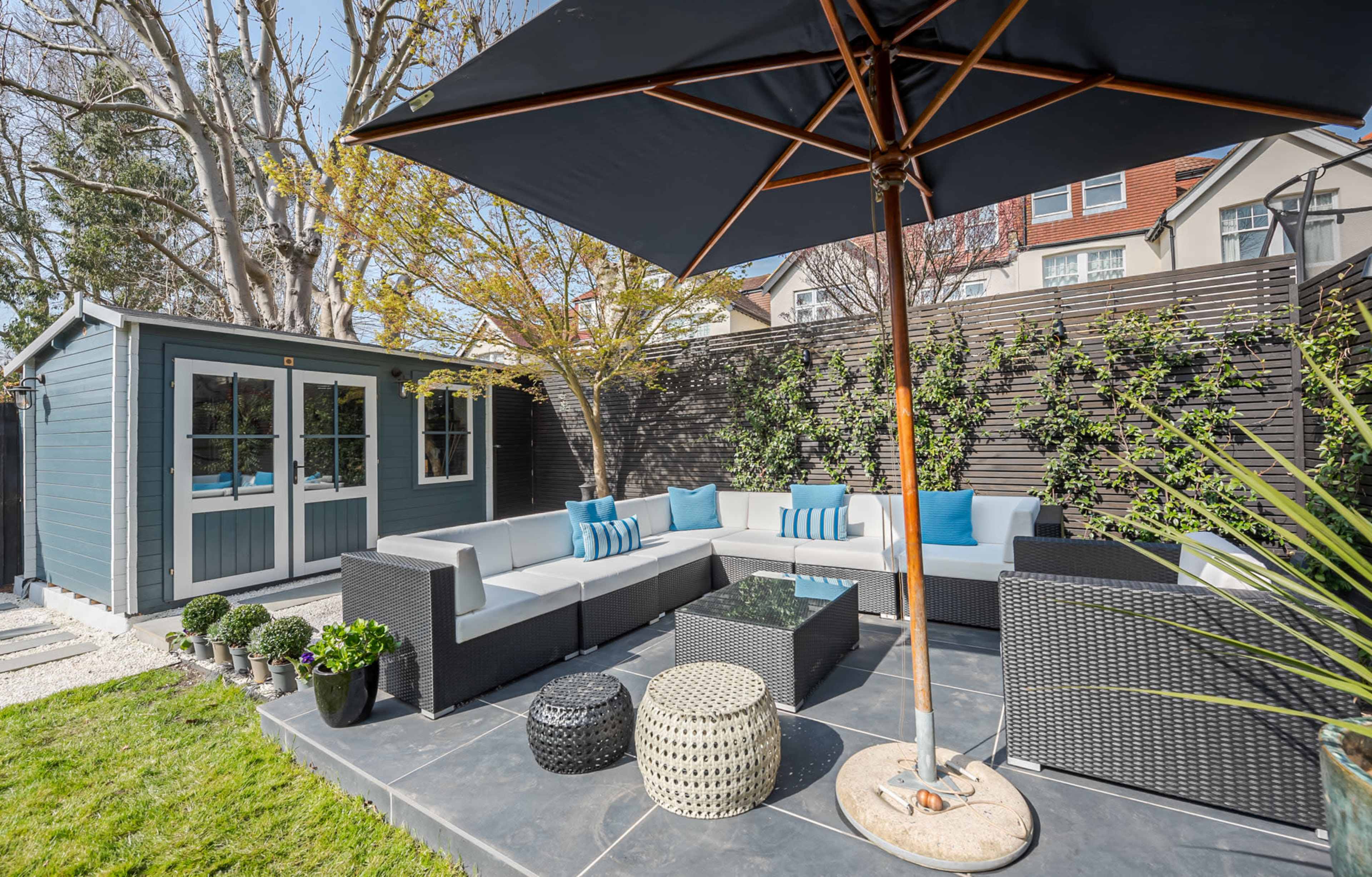 The image depicts a cozy outdoor seating area with a sectional sofa, an umbrella, and a garden shed surrounded by greenery.