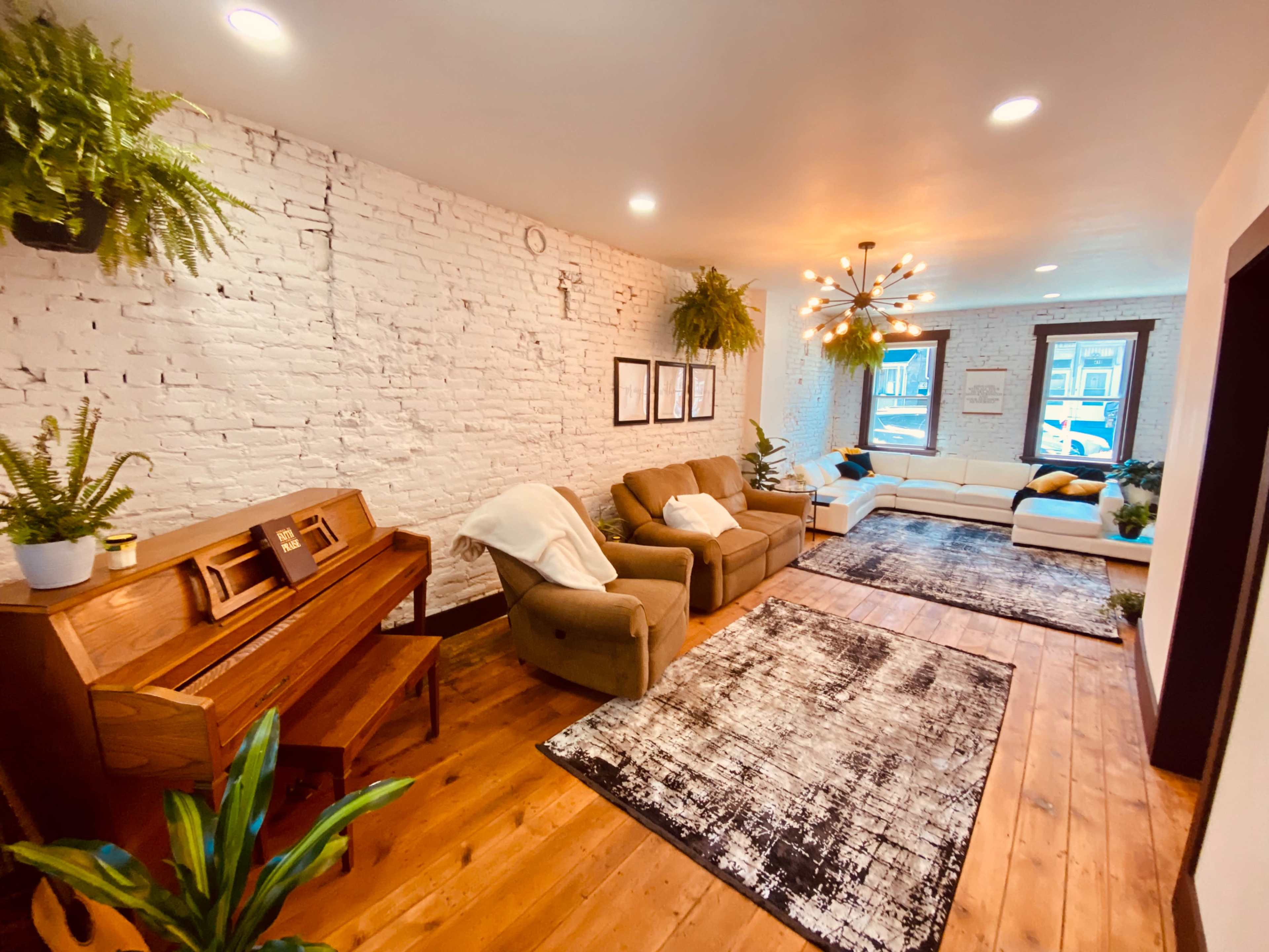 A spacious living room with white brick walls, wooden flooring, two couches, a piano, and plants hanging from the ceiling.