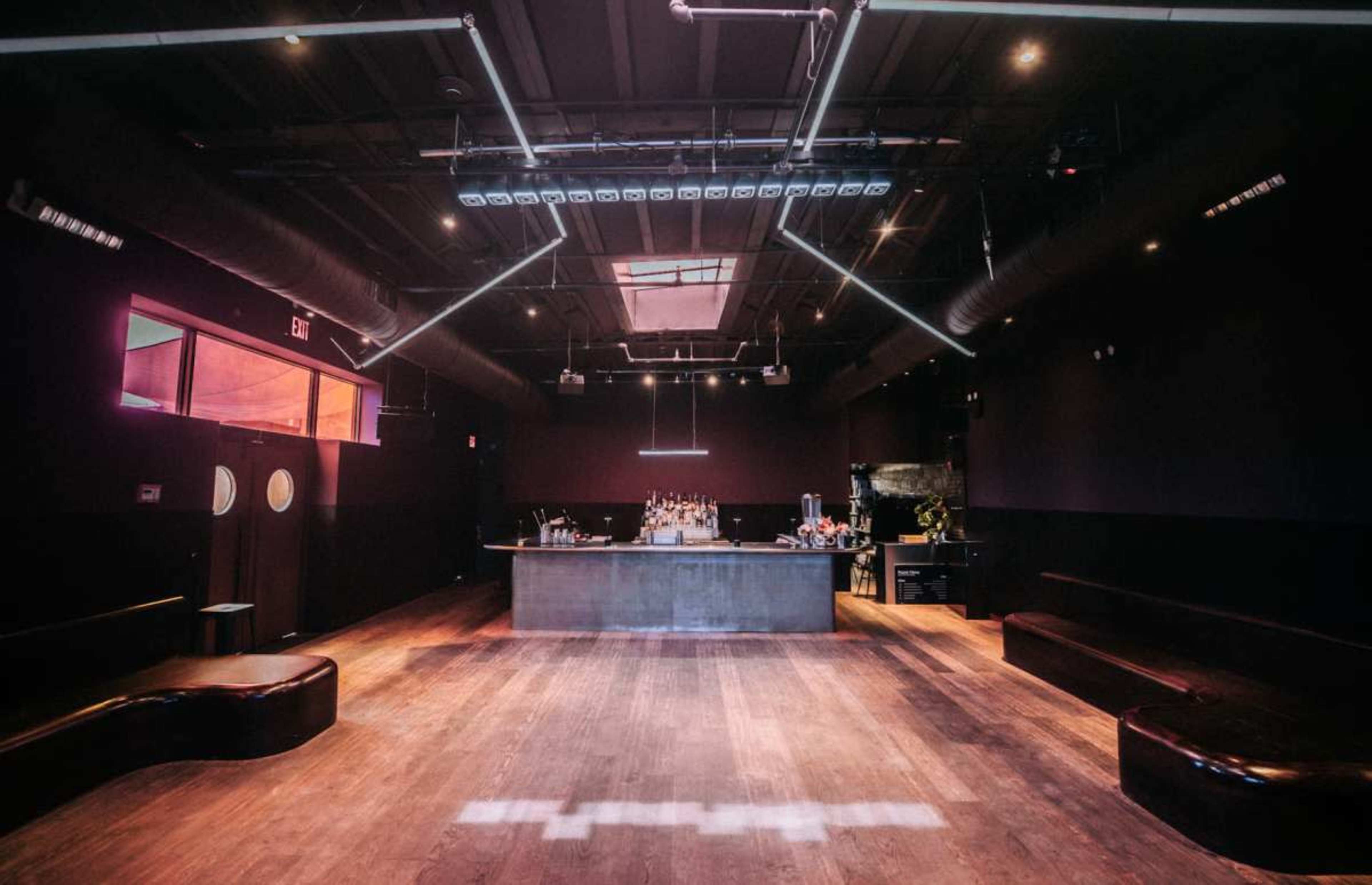 The image shows a dimly lit bar area with a wooden floor, sleek bar counter, and seating along the walls.