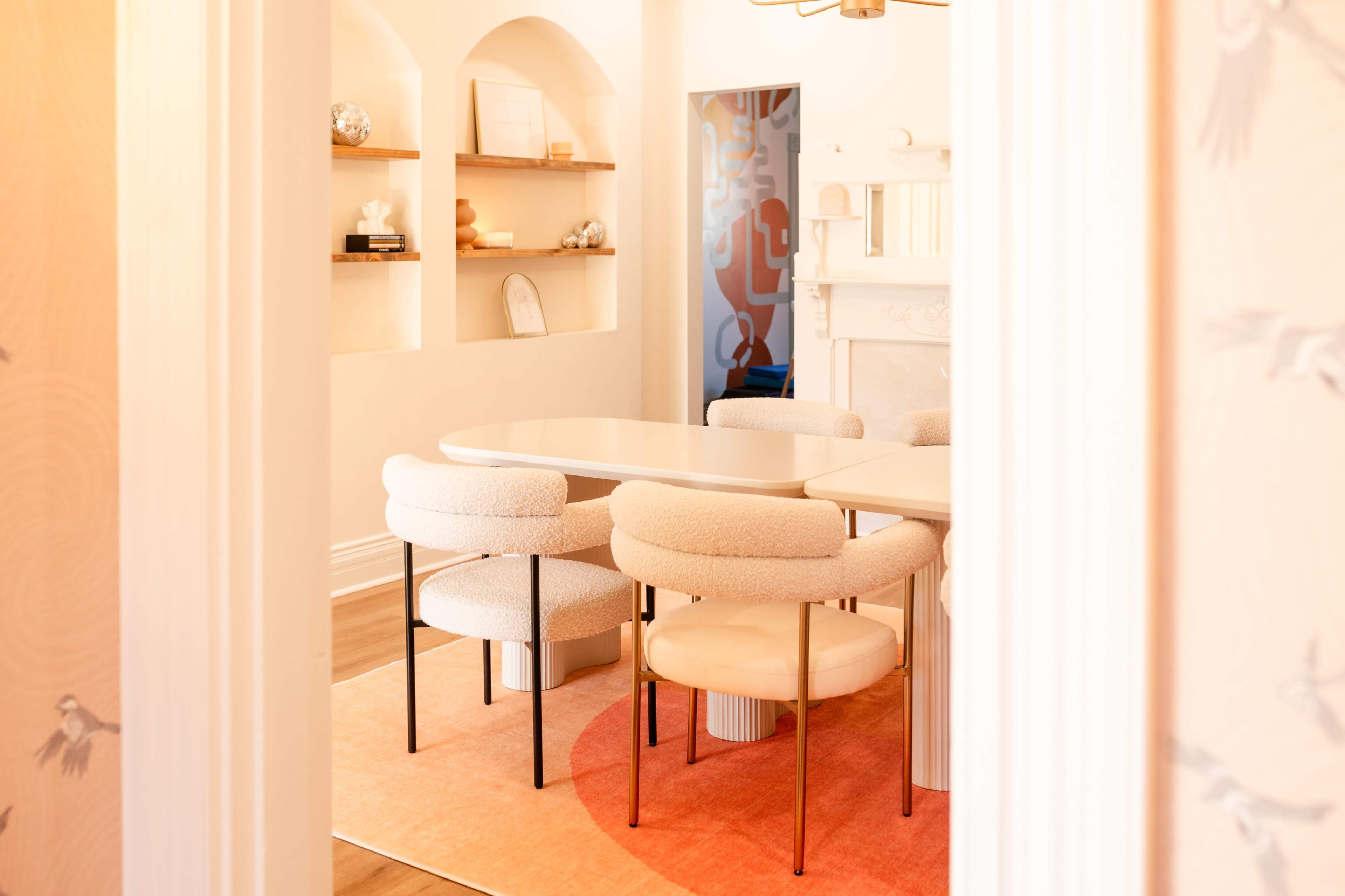 A modern dining area features a white oval table surrounded by plush chairs on a coral-colored rug, with shelves displaying decorative items in the background.