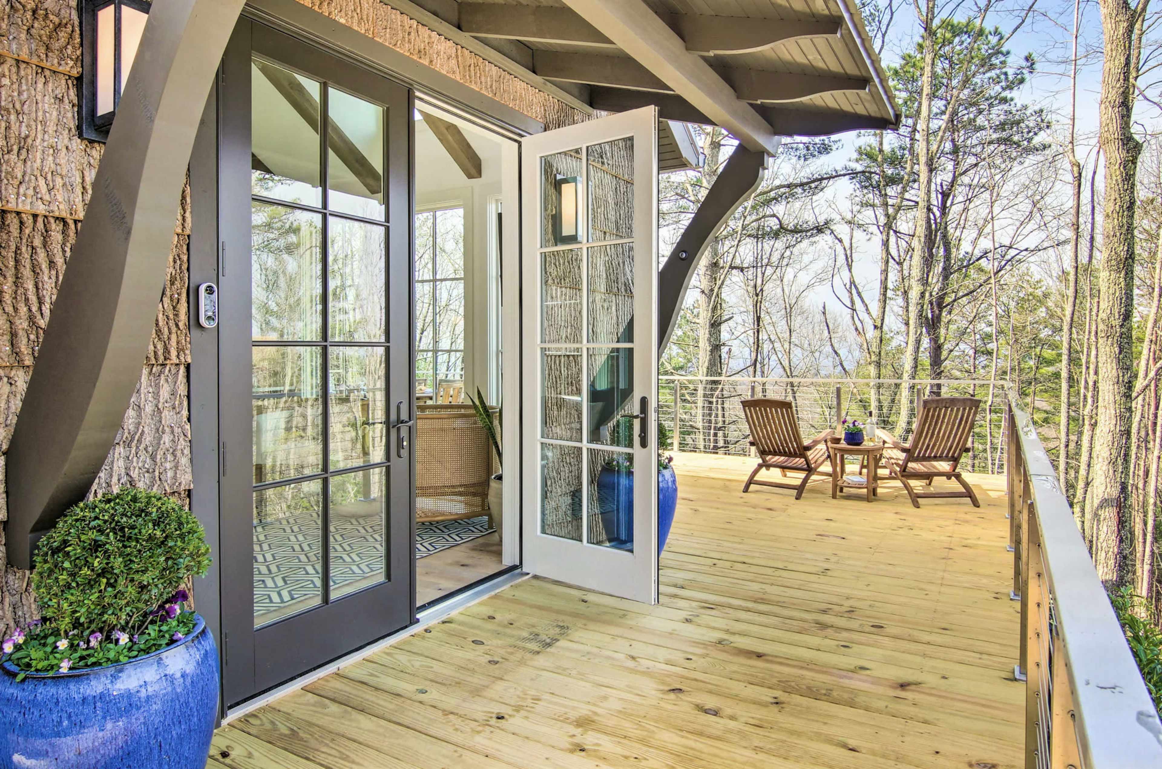 A wooden deck with two lounge chairs and a small table, accessed through glass double doors from a house, surrounded by trees.