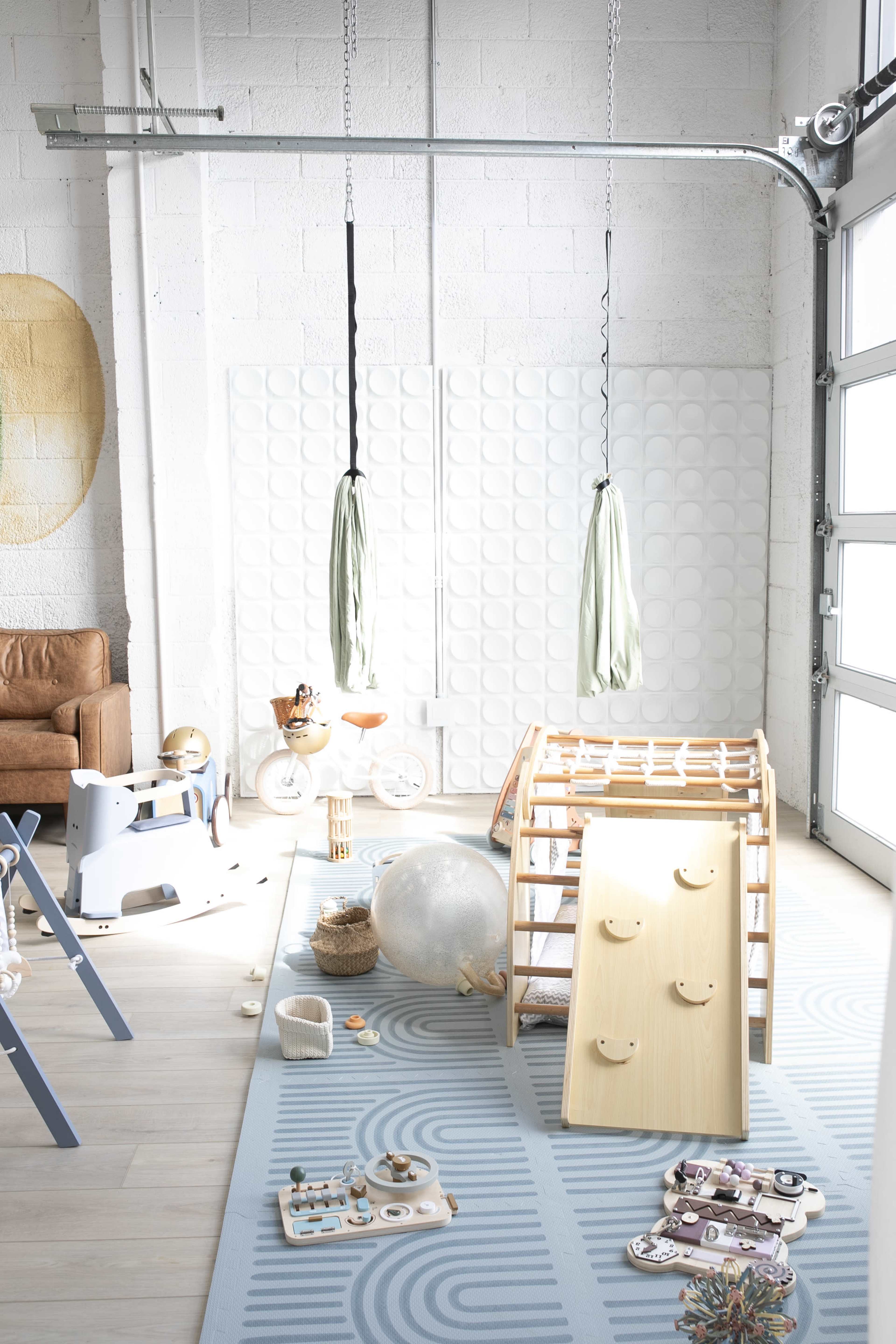 The image shows a bright and airy playroom featuring a wooden climbing frame, various toys scattered on the floor, and two hanging green fabric swings.