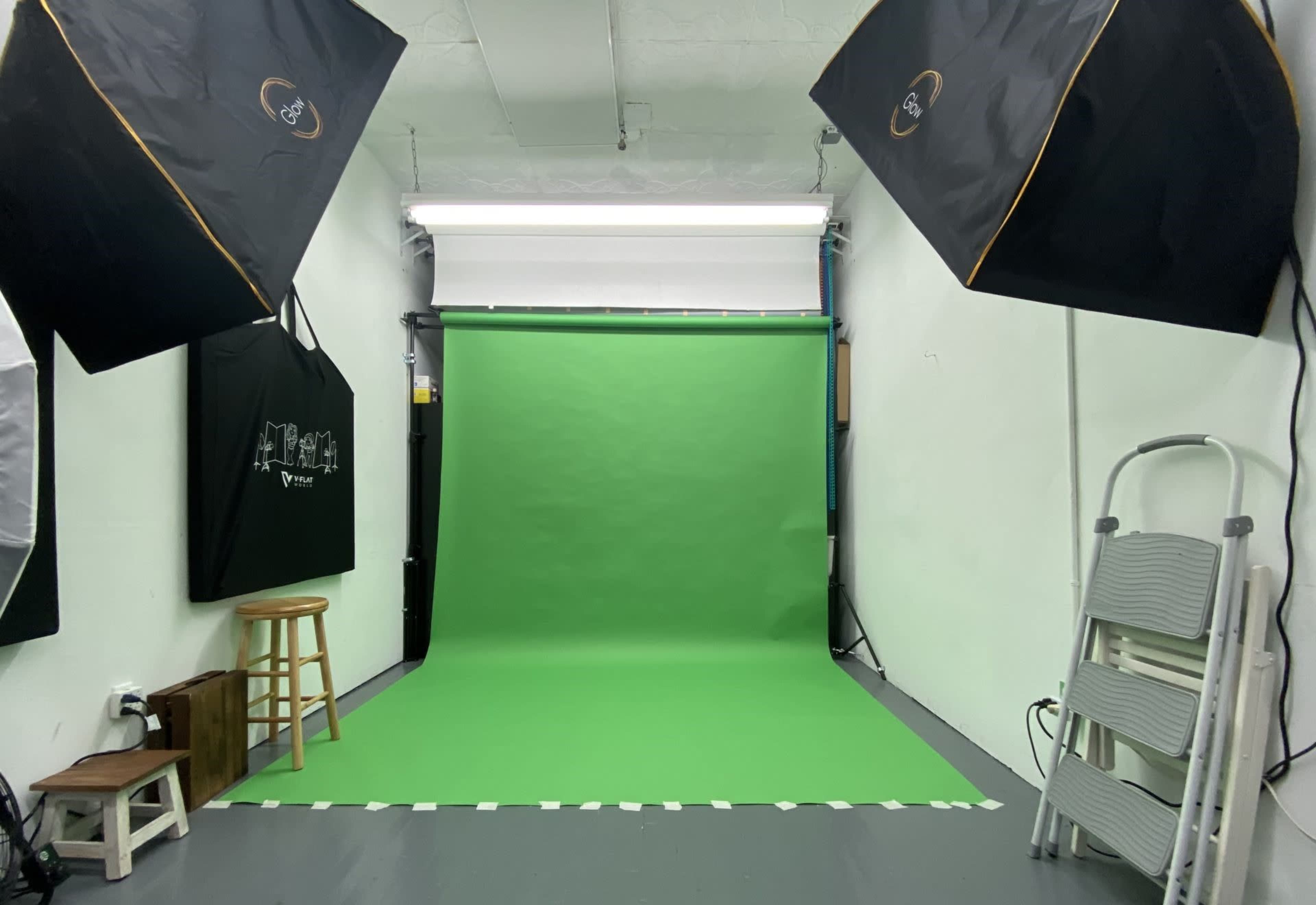 The image shows a photography studio featuring a green backdrop, two large softbox lights, and various items including a stool and a folding ladder.