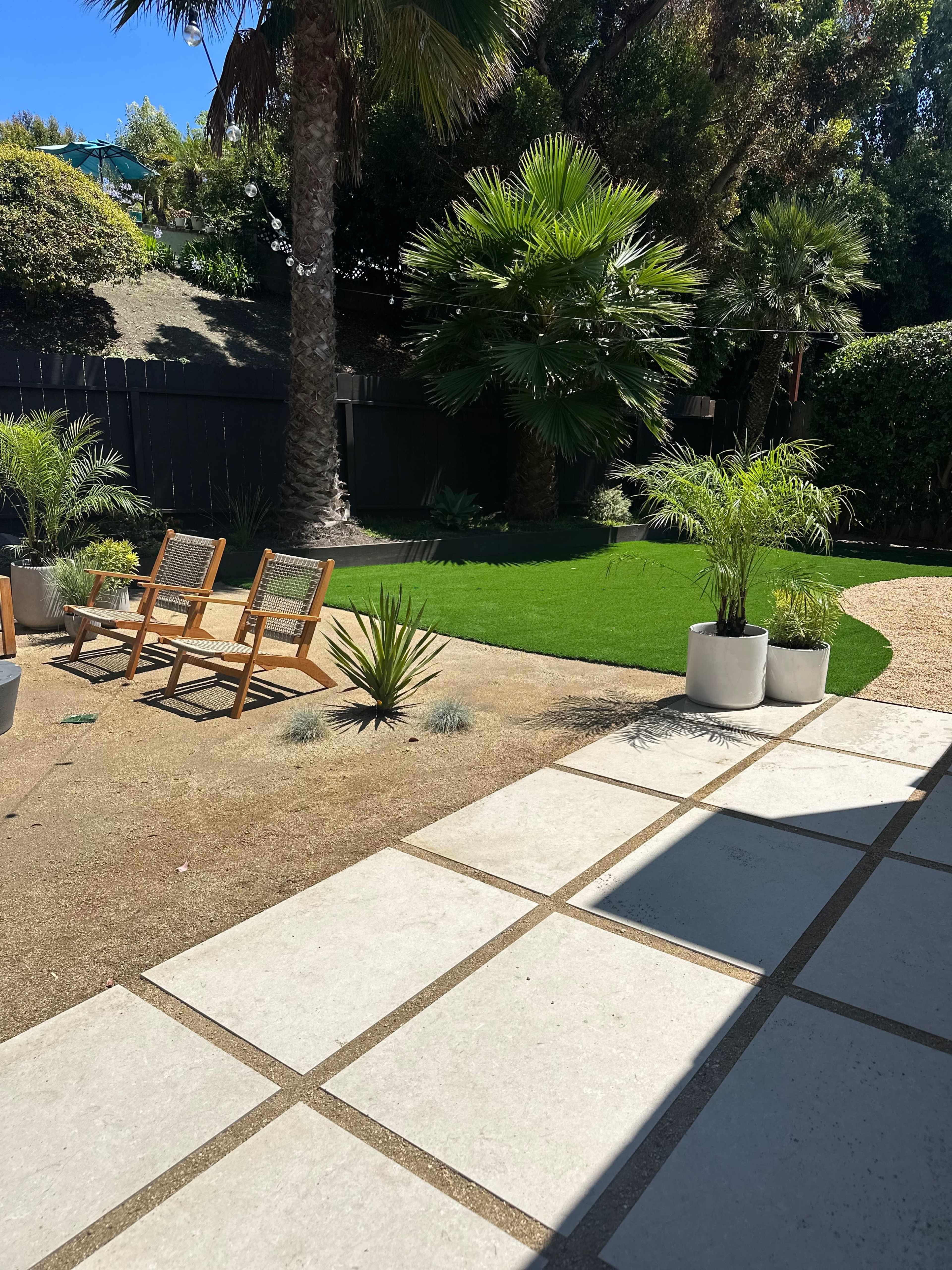 A landscaped outdoor area featuring two wooden chairs, artificial grass, potted plants, and a stone pathway bordered by gravel.