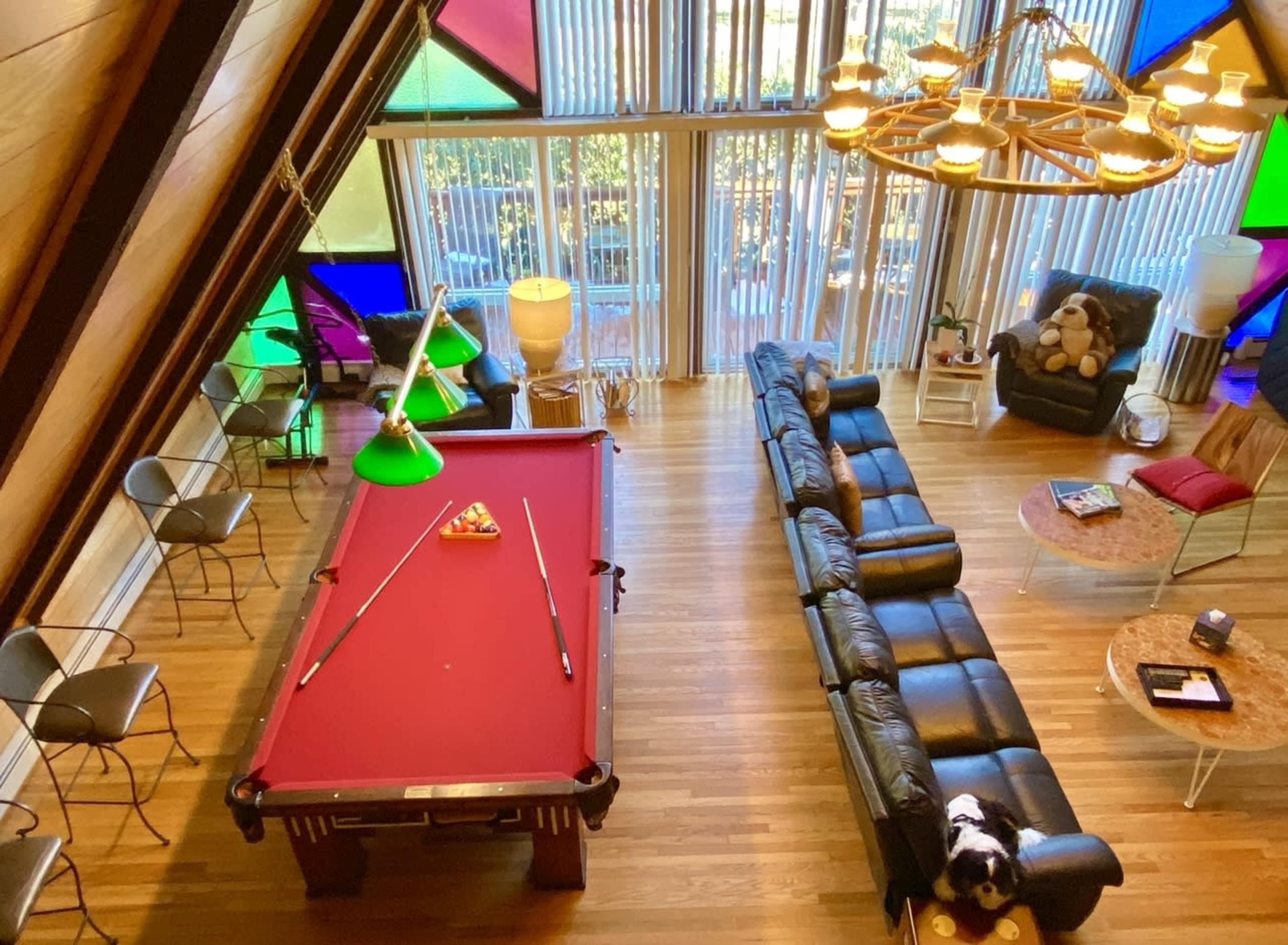The image shows a spacious room with a pool table, a black leather sectional sofa, and large windows featuring colorful glass panels.