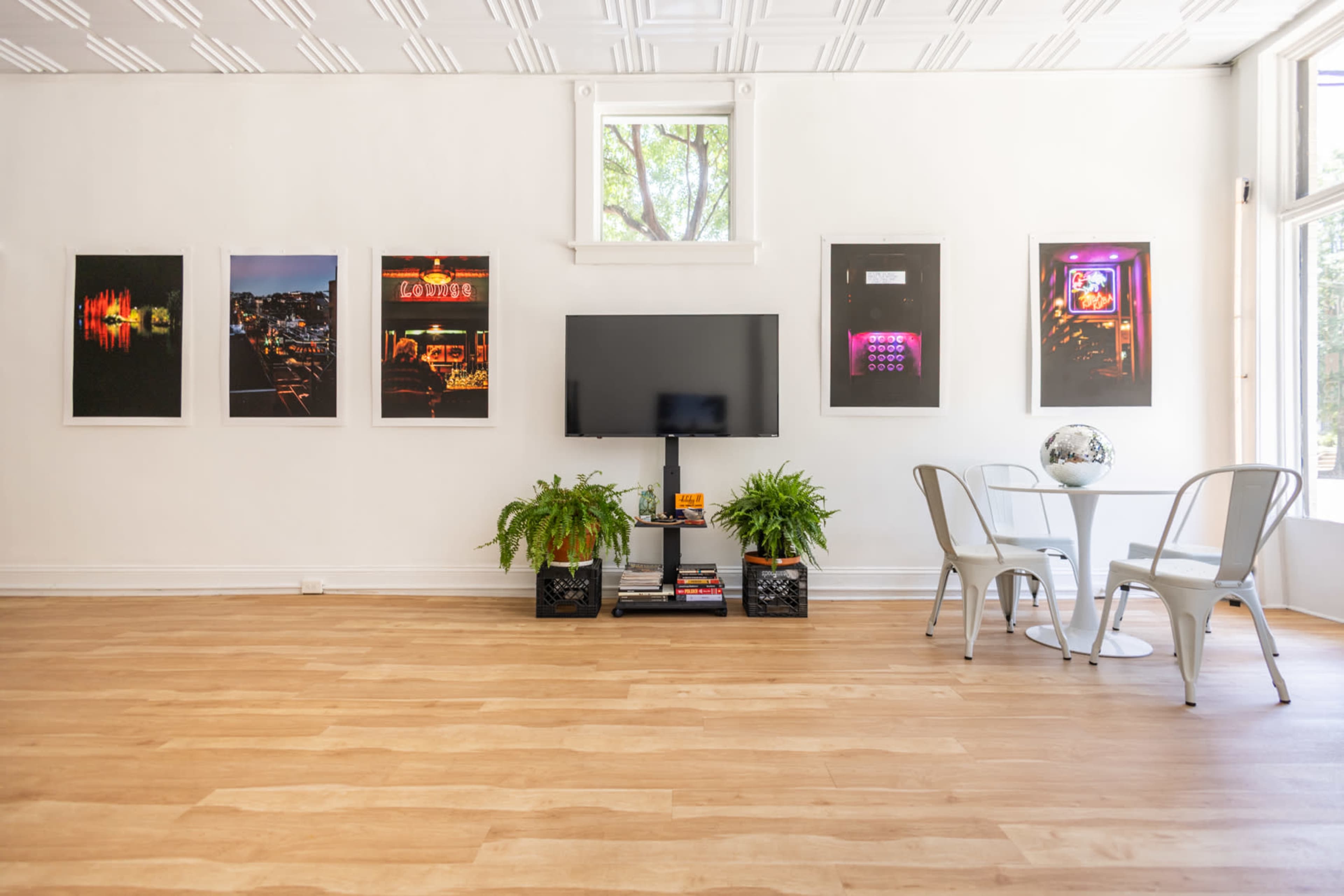 The image shows a bright room with wooden flooring, featuring a wall adorned with framed photographs, a television mounted on the wall, and a small dining table with two metal chairs.