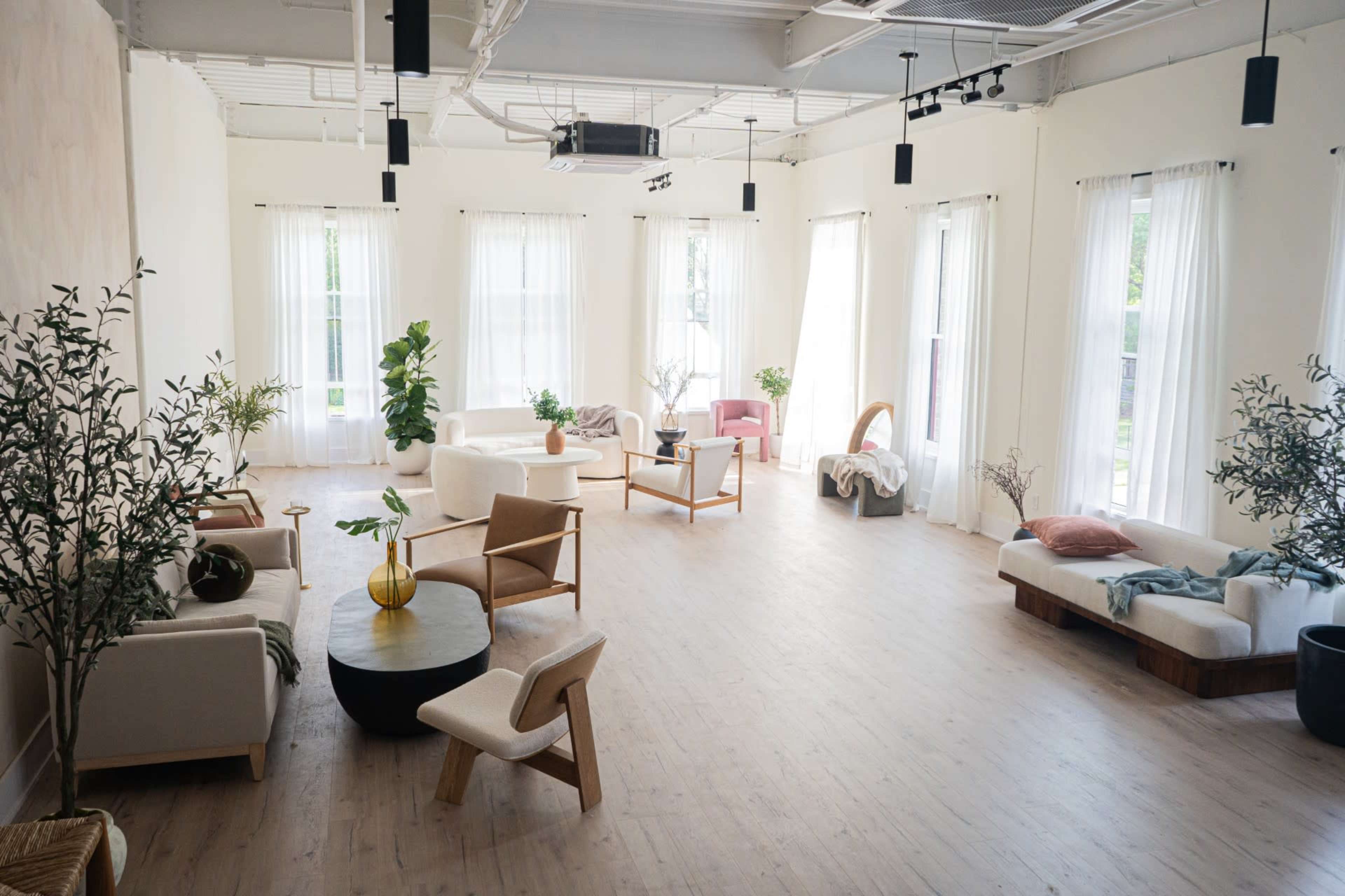 A spacious, well-lit room features several modern seating arrangements amid tall windows and decorative plants.