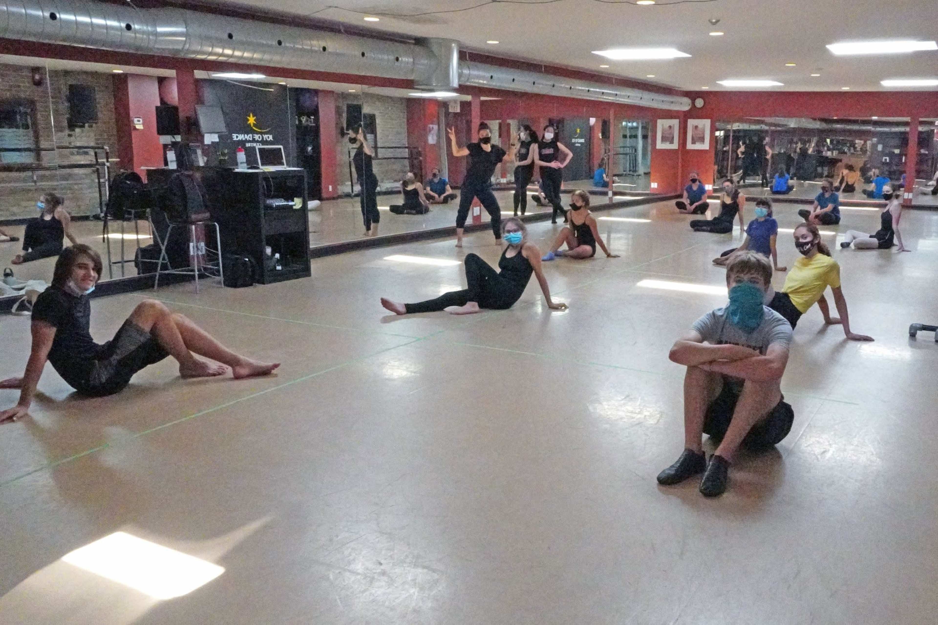 A group of dancers wearing masks is seated on the floor of a studio, with some standing and stretching in the background.