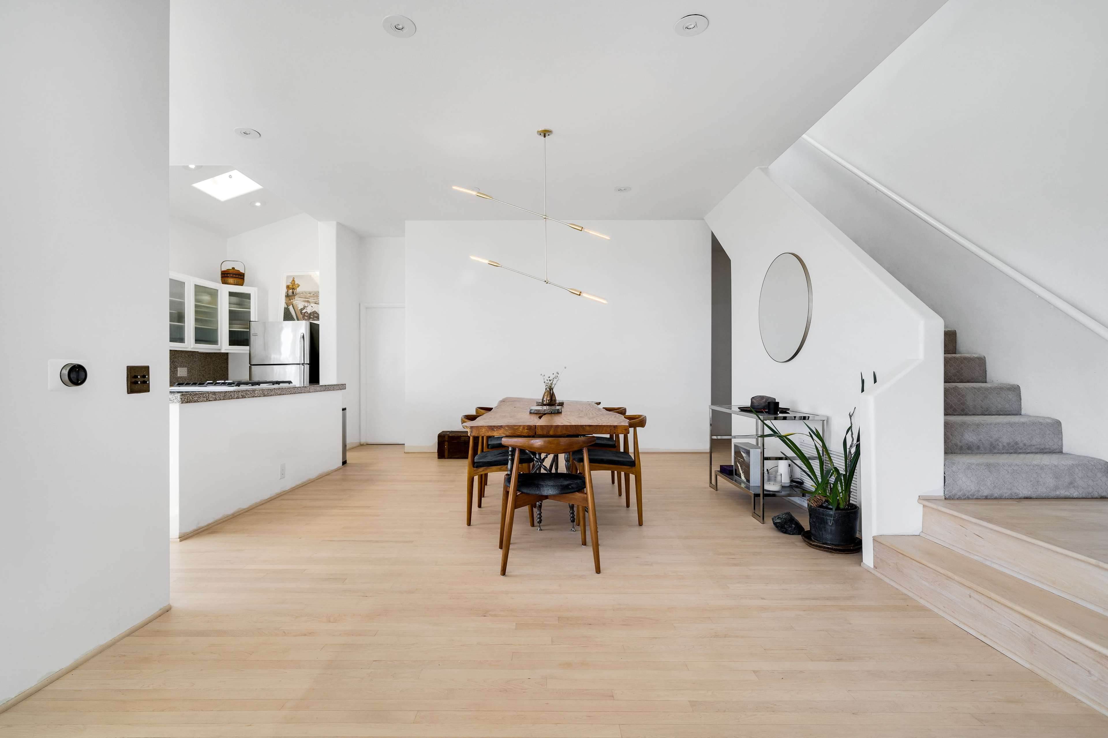 A modern dining area features a long wooden table surrounded by chairs, with an open kitchen visible in the background and a staircase leading to an upper floor.
