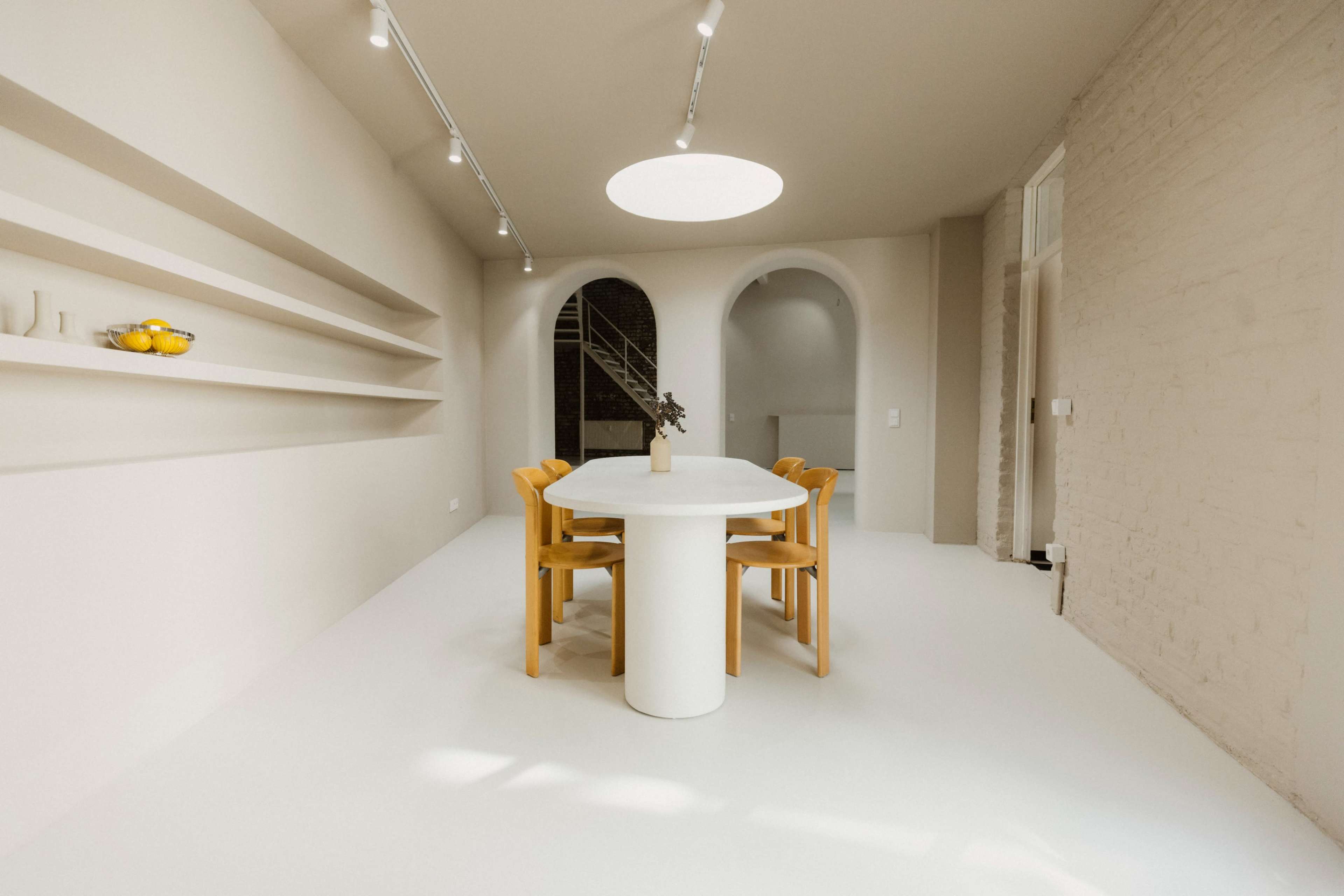 A minimalistic dining area features a round table surrounded by wooden chairs, with shelves displaying simple decor and a soft, neutral color palette.