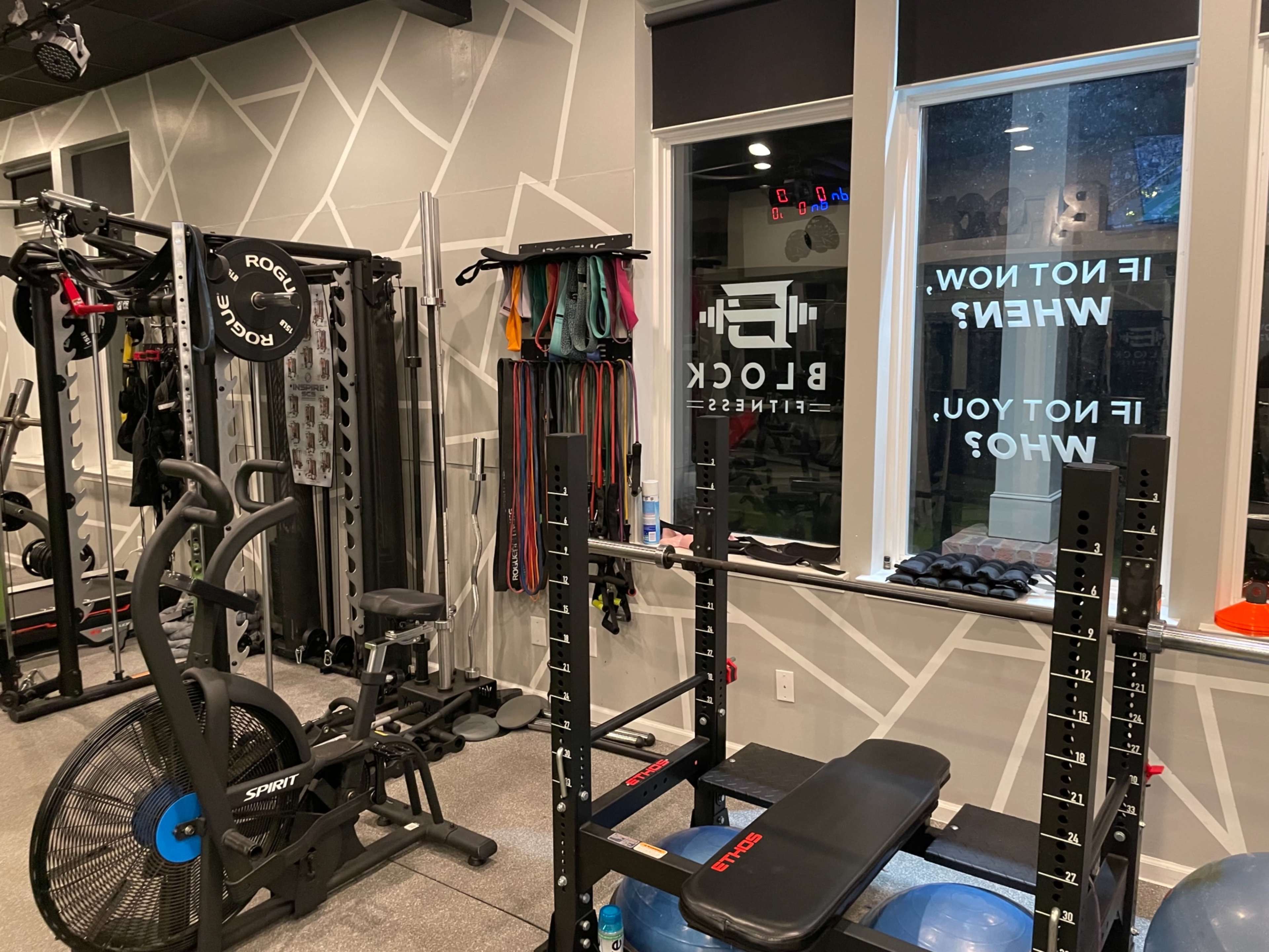 A fitness studio equipped with exercise machines, weights, and resistance bands, featuring large windows with motivational phrases.