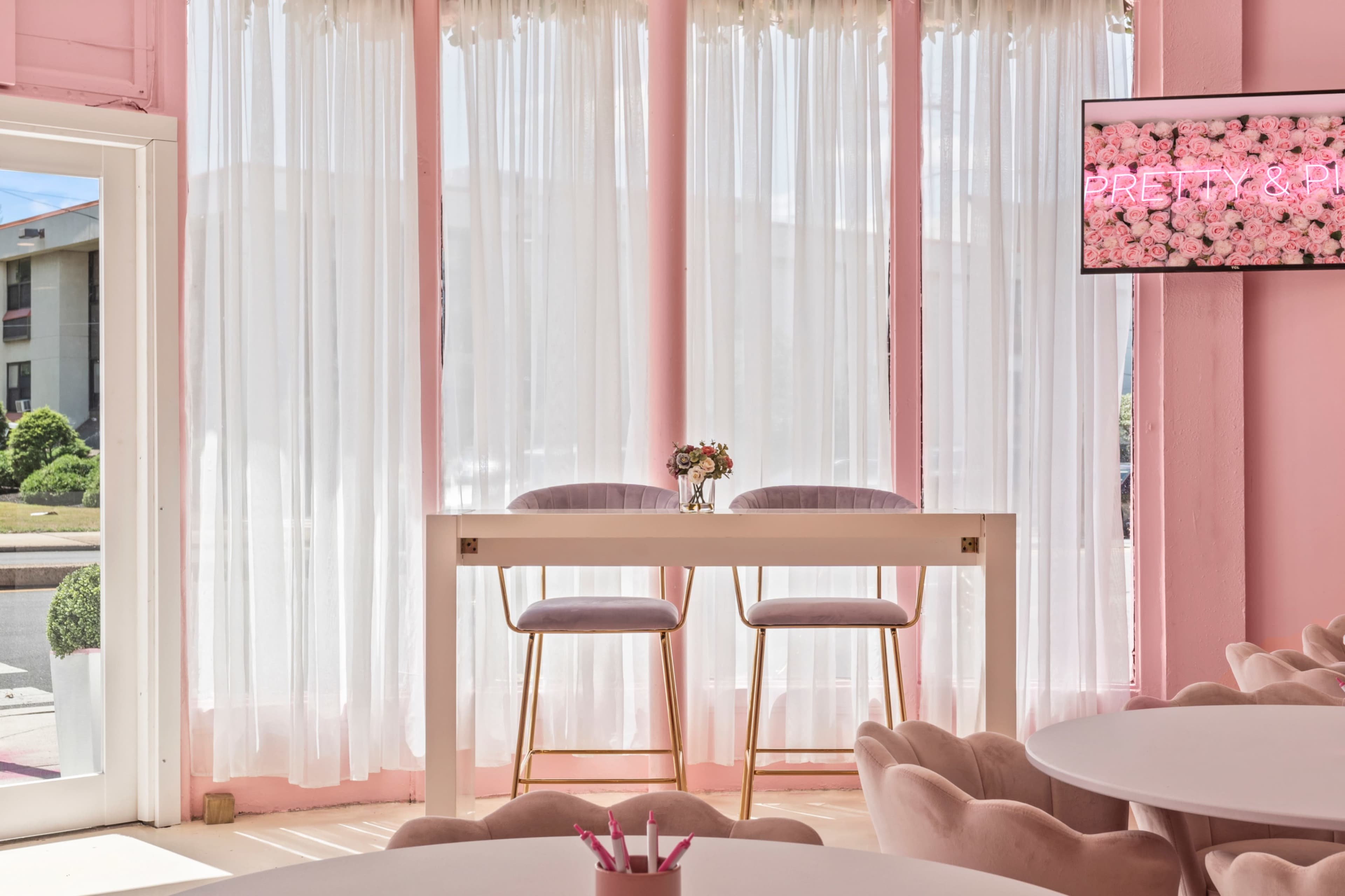 The scene features a bright interior with large sheer curtains, a white table with two stools, and a television displaying a floral-themed text.