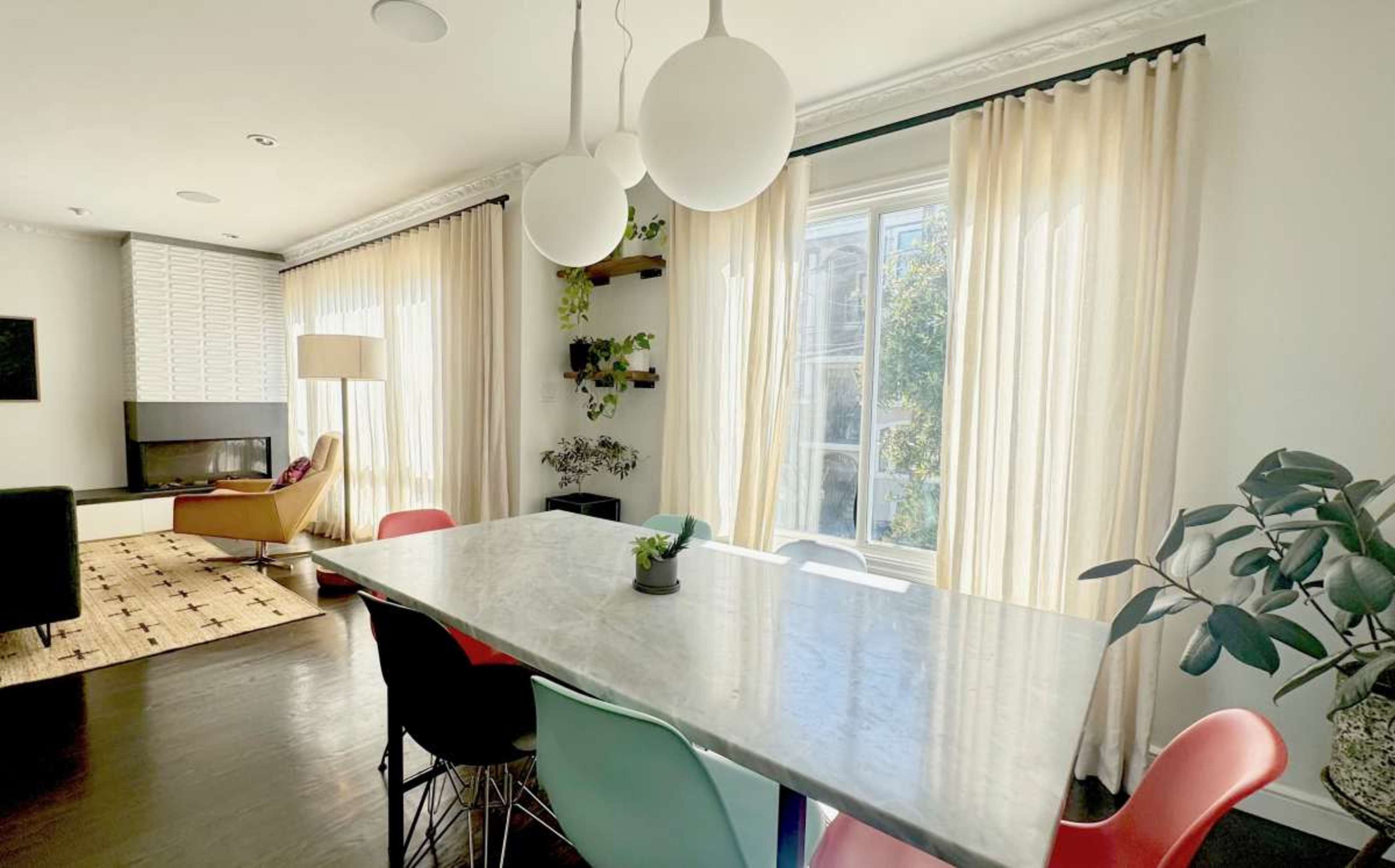 The image shows a modern living space featuring a dining table with colorful chairs, large windows with sheer curtains, and a combination of indoor plants and contemporary furniture.