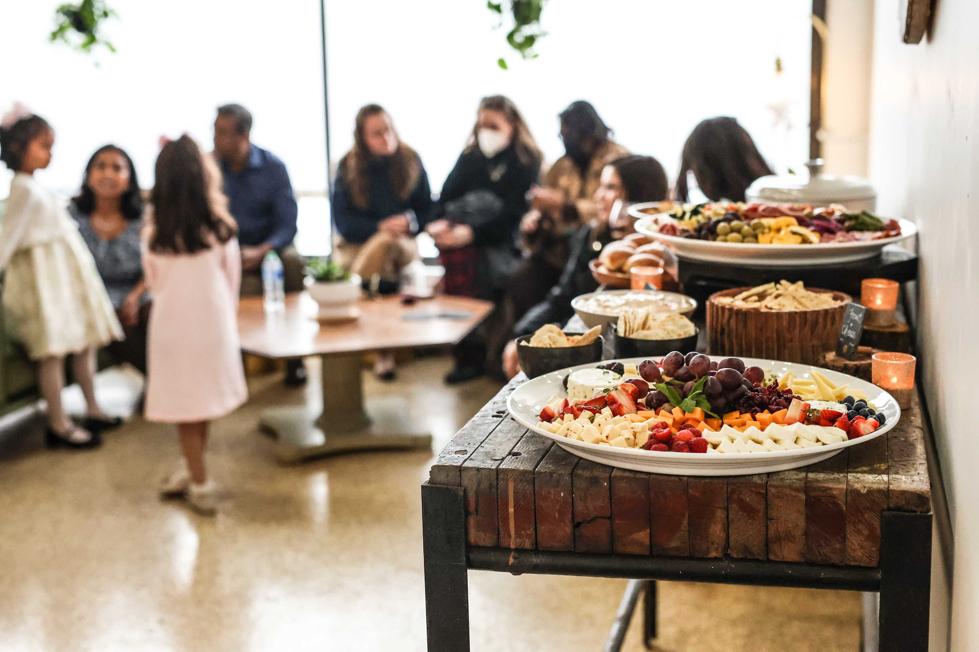 A table displays an assortment of fruit and cheese platters while a group of people sits in the background.