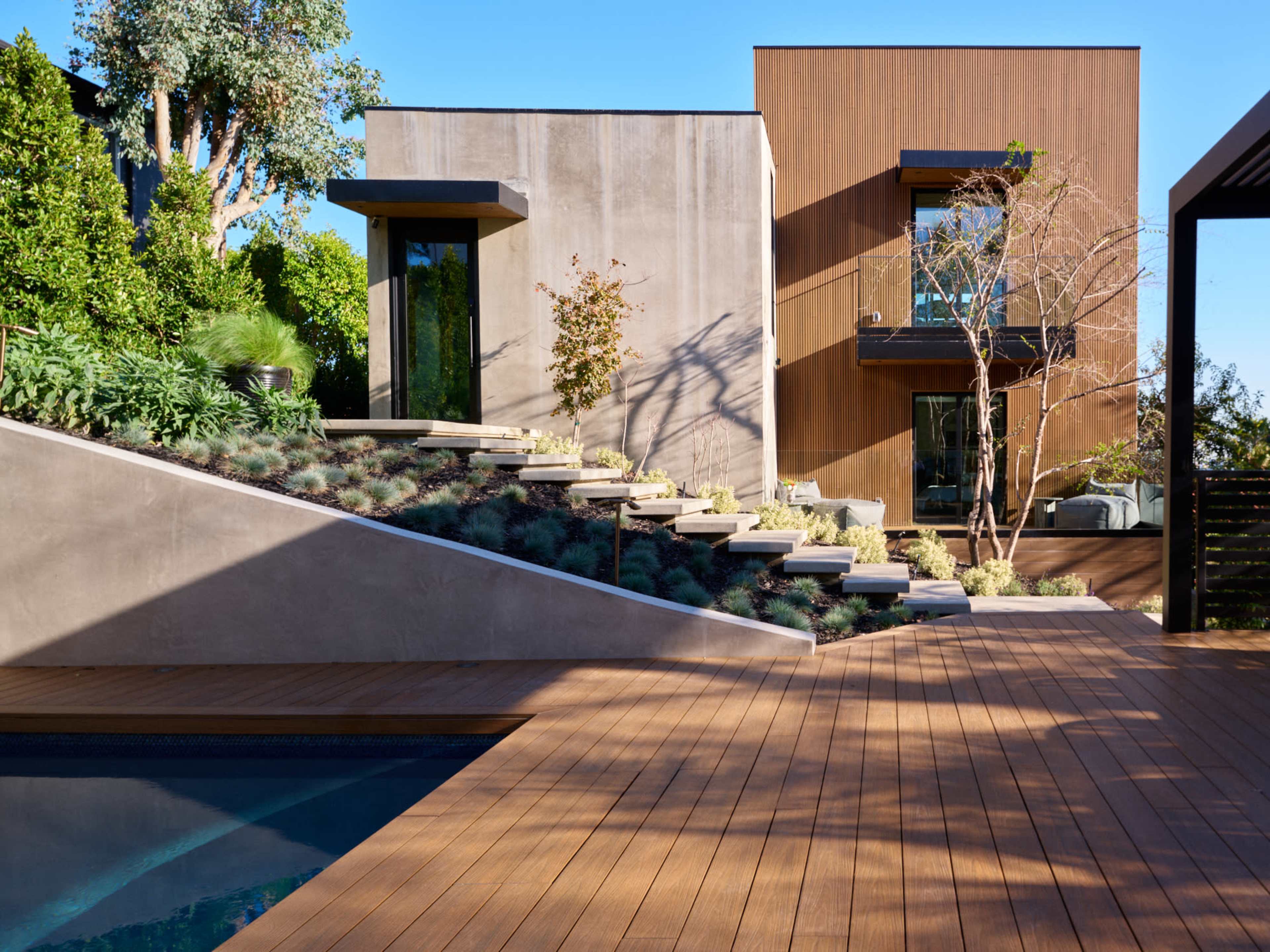 The image depicts a modern home with two distinct sections, a landscaped staircase, and a wooden deck surrounding a pool.