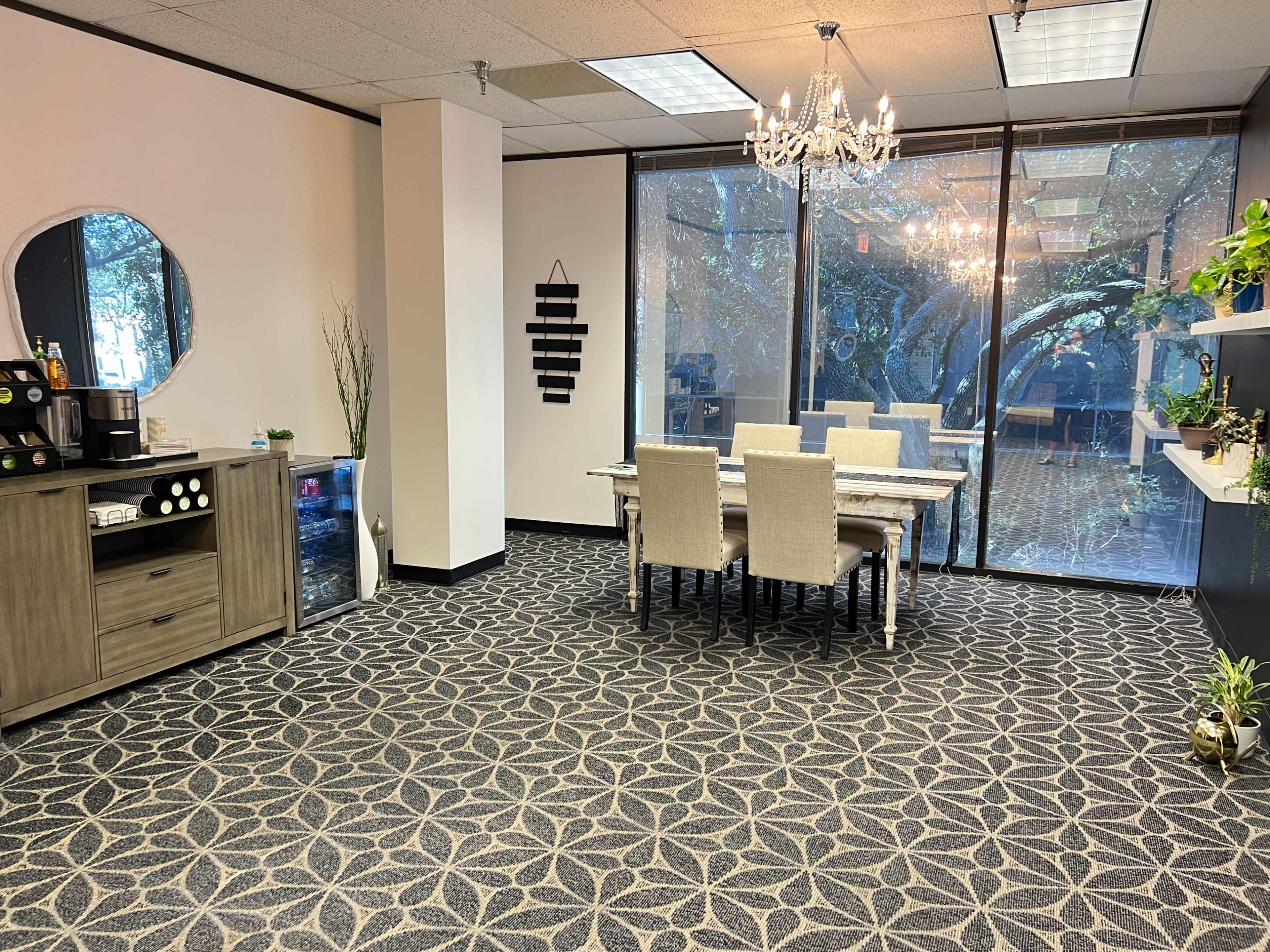 A modern office space featuring a dining table set for six, a coffee area with a cabinet, and large windows with views of trees outside.