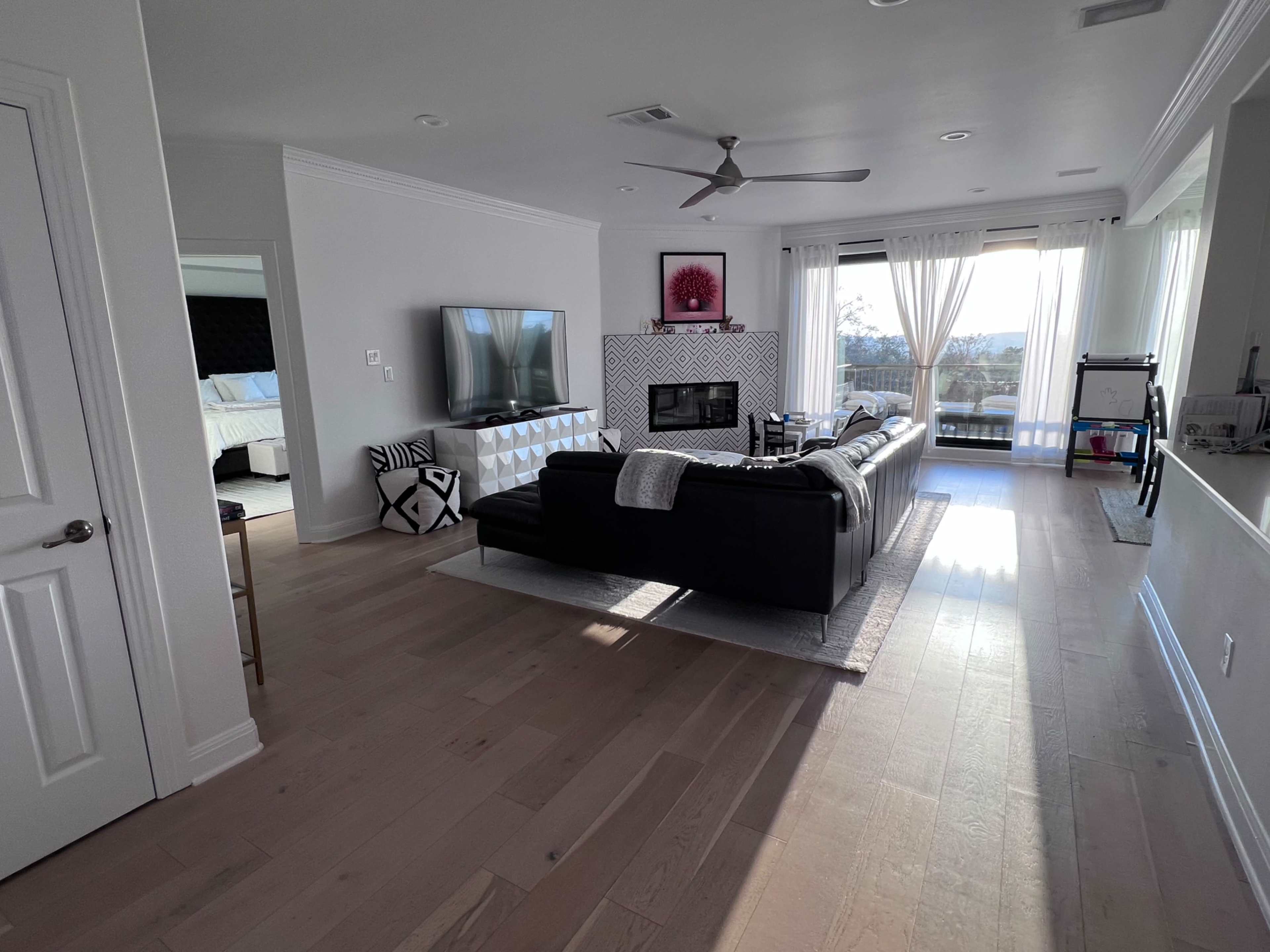 The image shows a spacious living room with a large black sofa, a wall-mounted TV, and large windows that offer a view of the outdoors.
