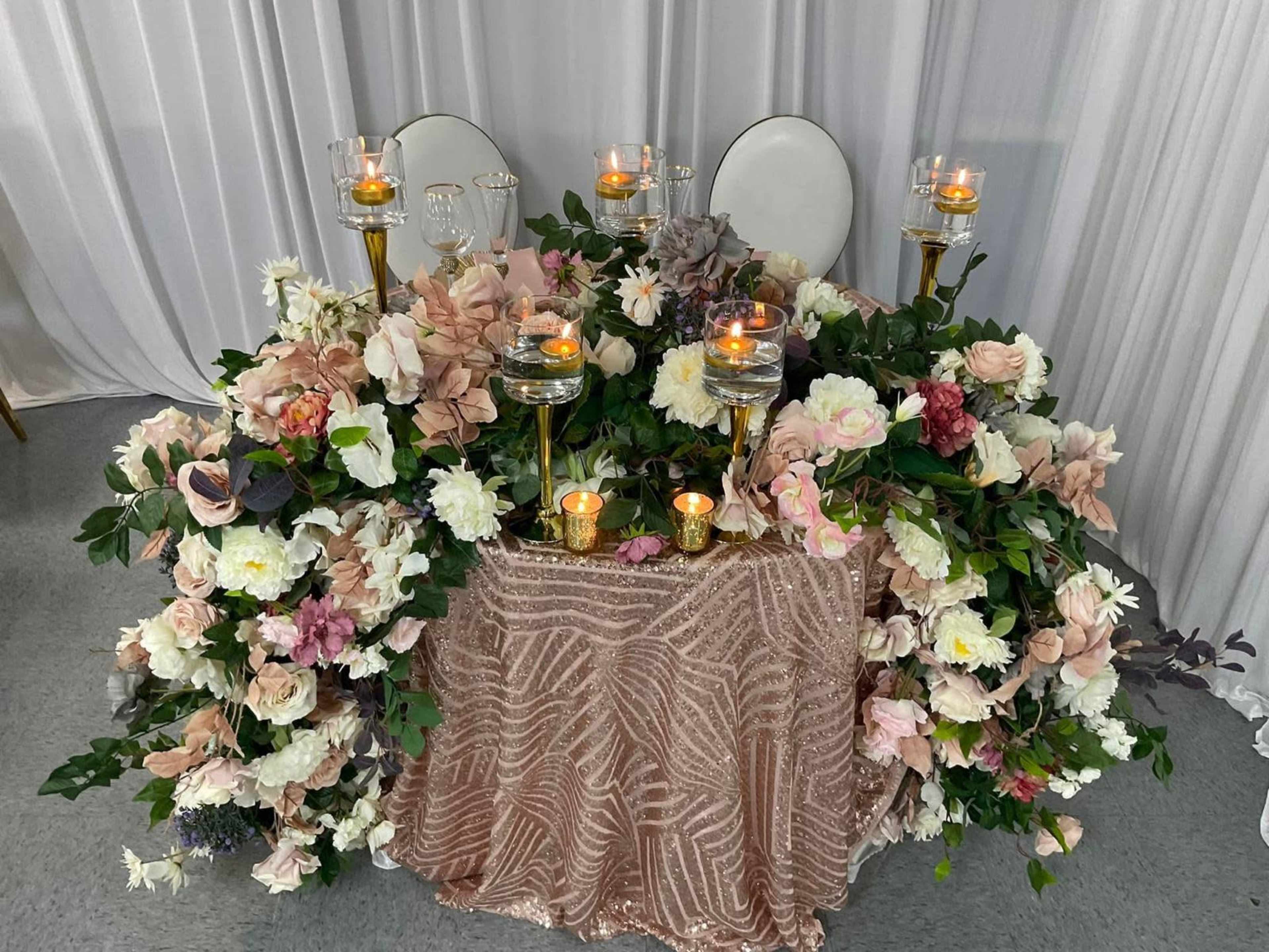 The image shows a floral-decorated table with candles and two empty chairs in front of a backdrop.