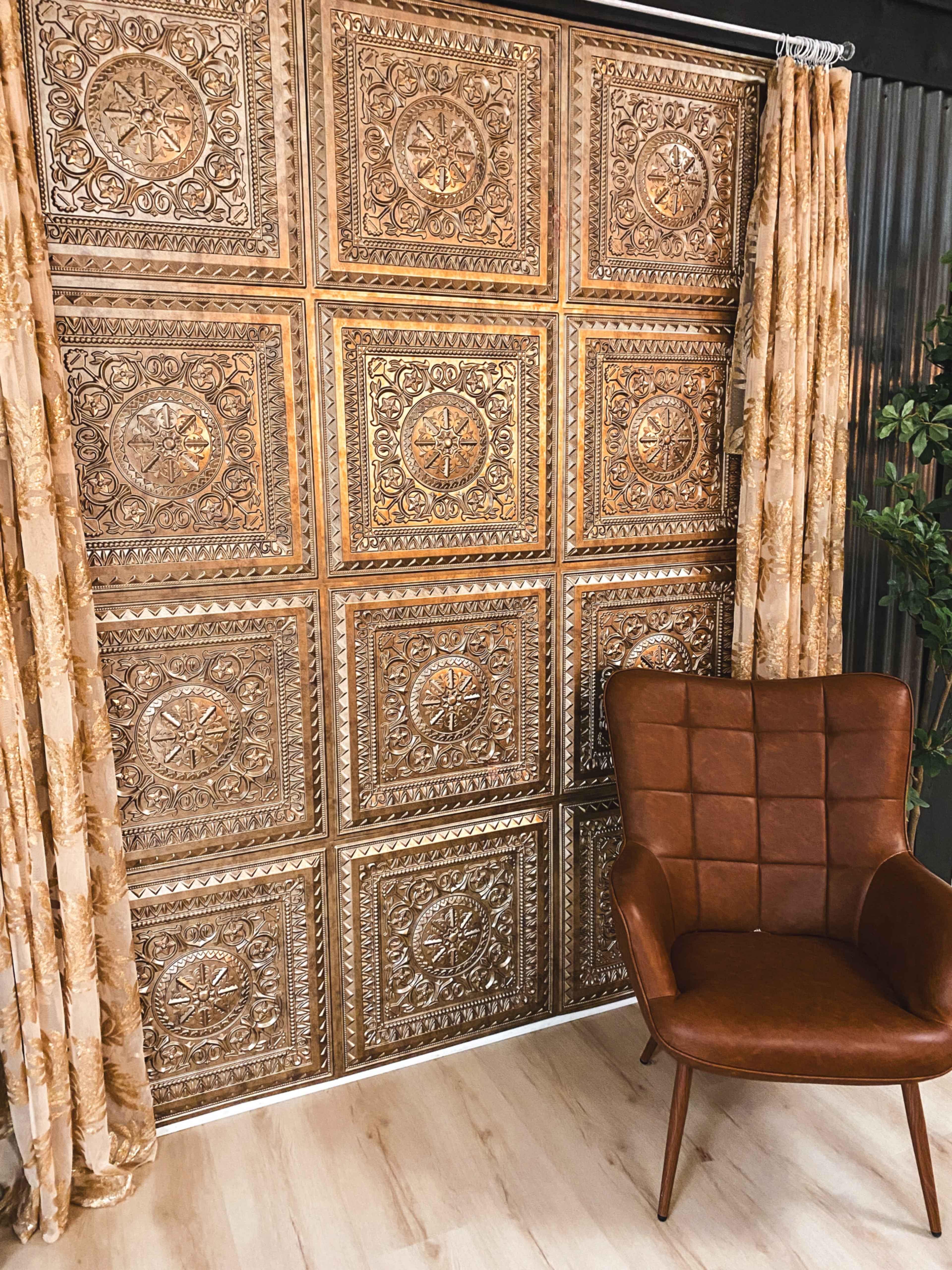 The image shows a decorative wall with a patterned metallic panel and a brown chair in front of it, accompanied by patterned curtains and a potted plant in the background.