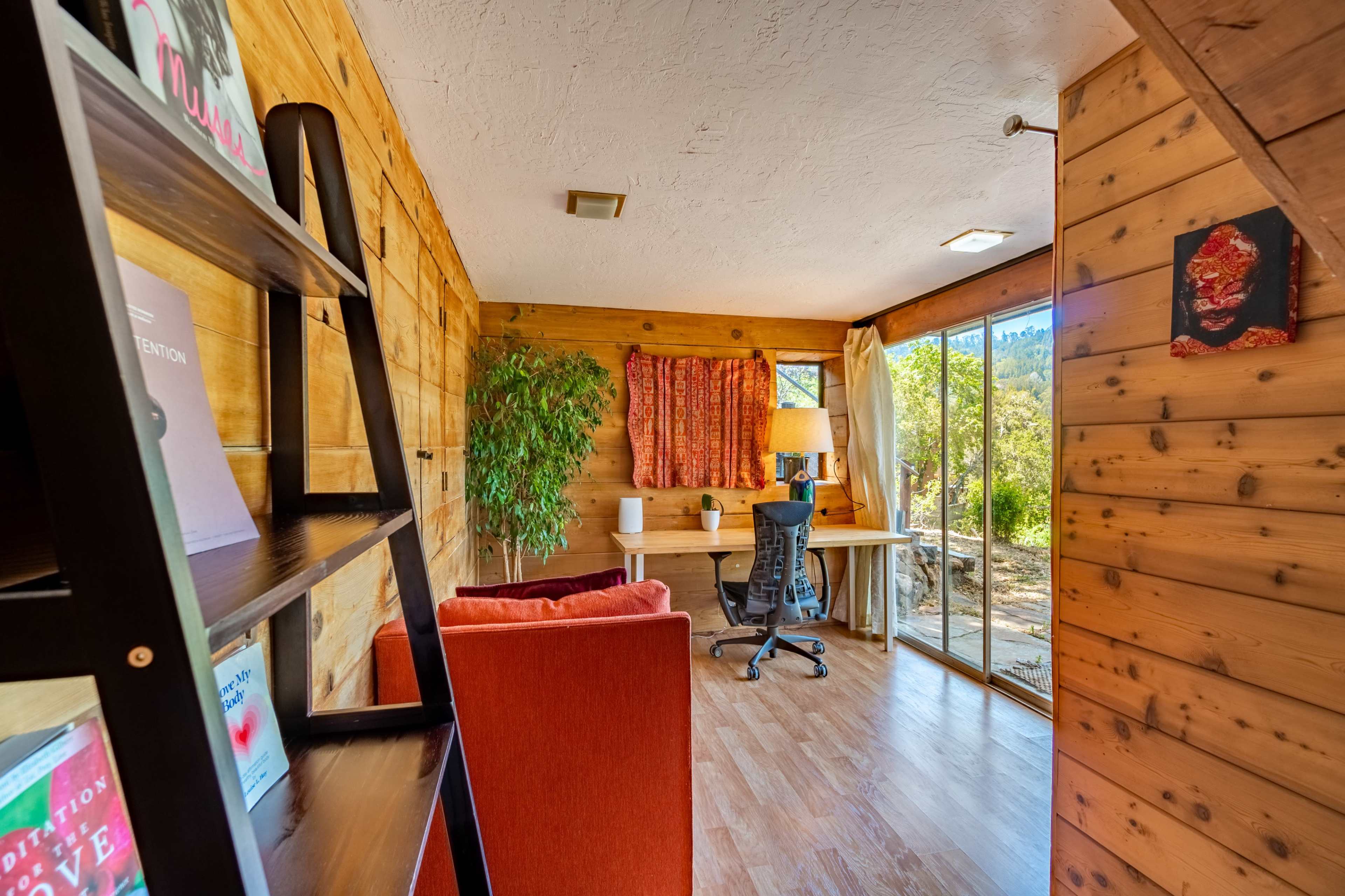 A small wooden room features a desk and chair next to a large window, with a bookcase and two red chairs nearby.