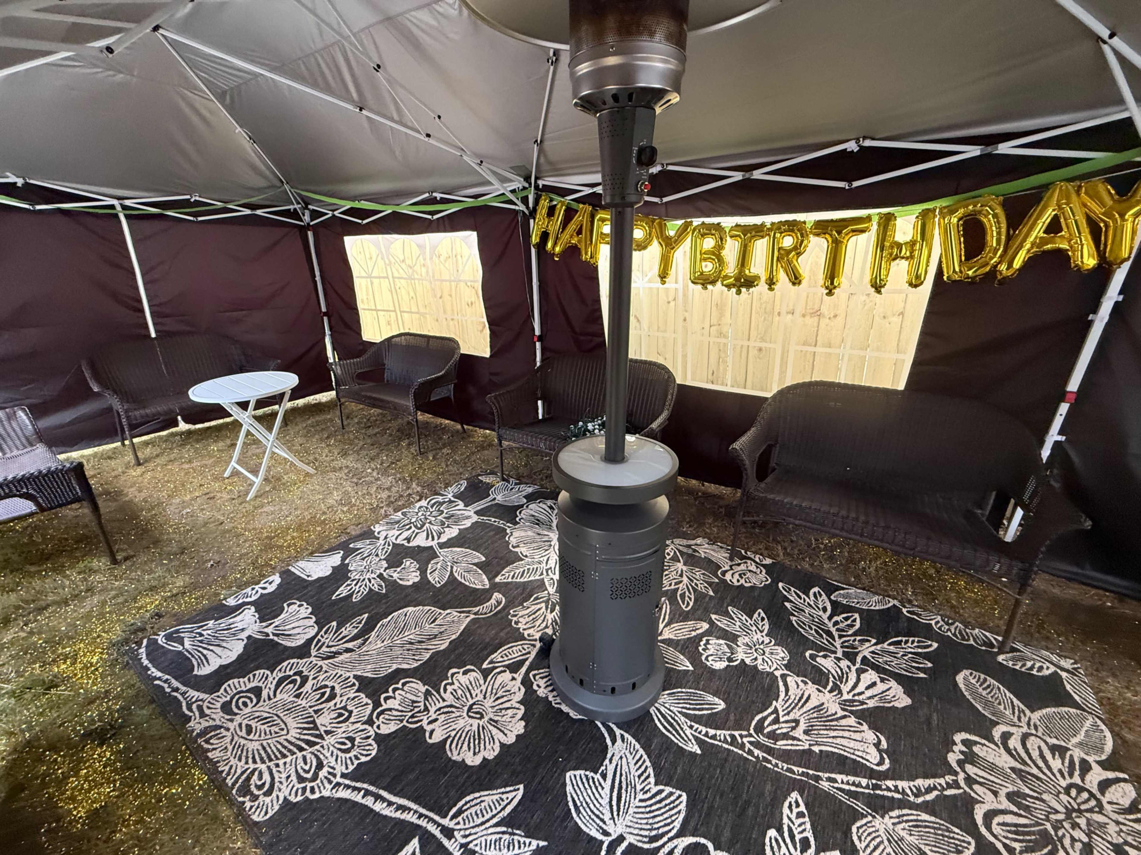 The image shows a tented area with a decorative "HAPPY BIRTHDAY" banner, a central heater, and seating arranged on a floral-patterned rug.