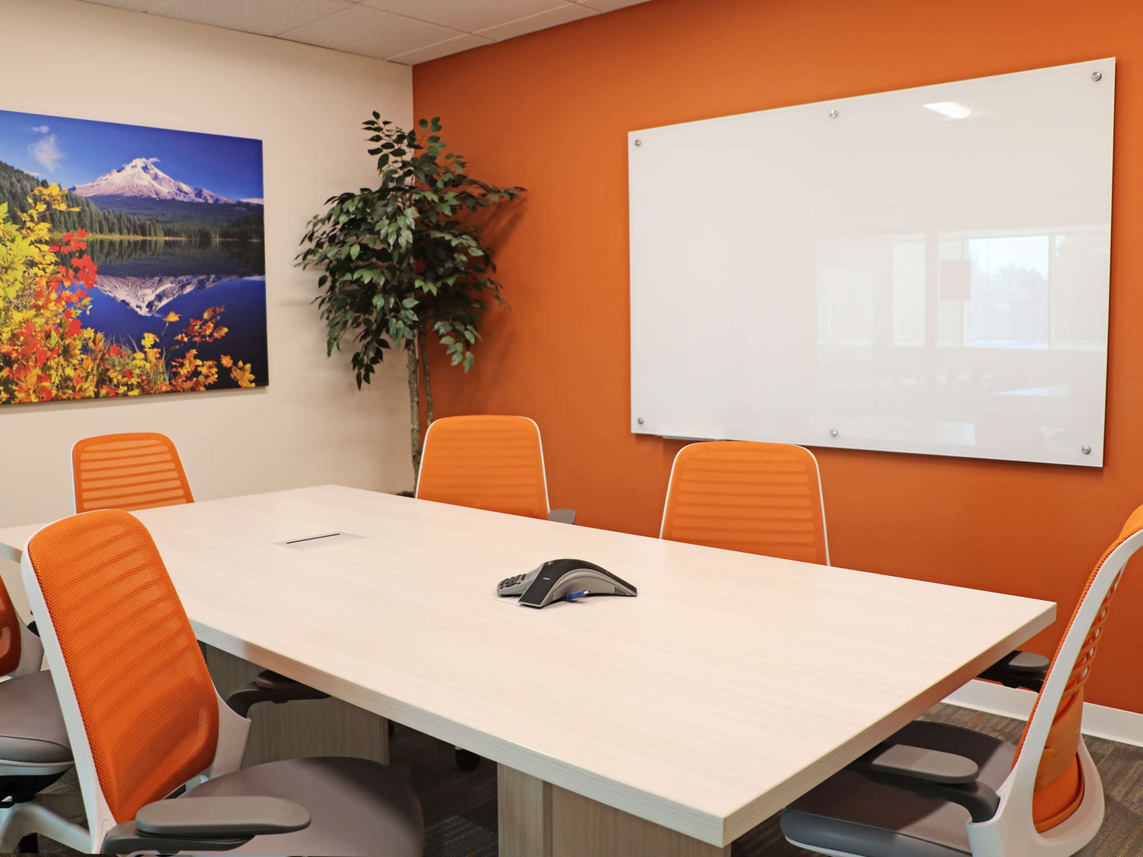 A conference room features a long table surrounded by orange chairs, a whiteboard on the wall, and a landscape painting depicting a mountain scene.