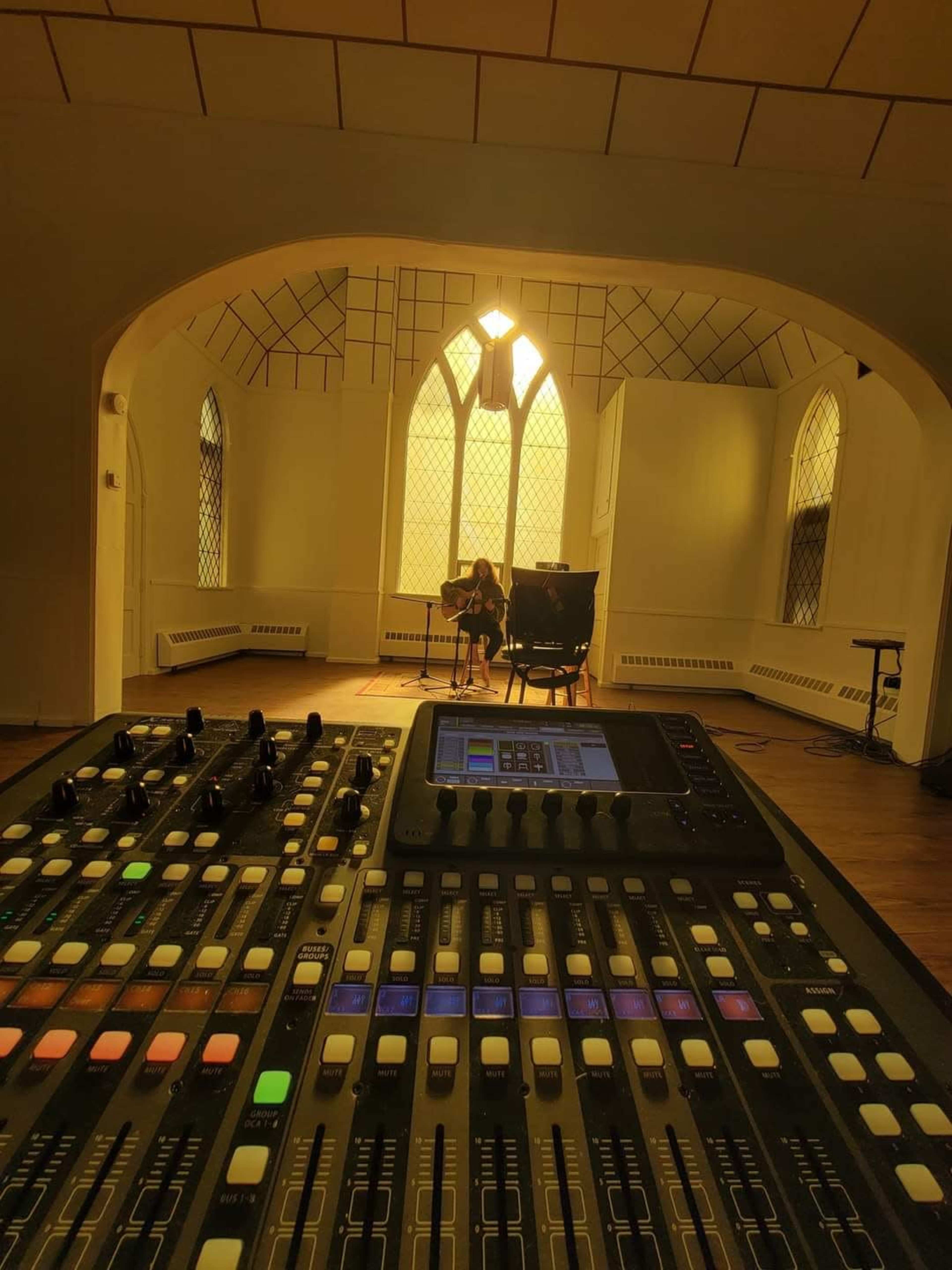 A musician plays guitar on a stage surrounded by soft, golden light in a room with large windows.