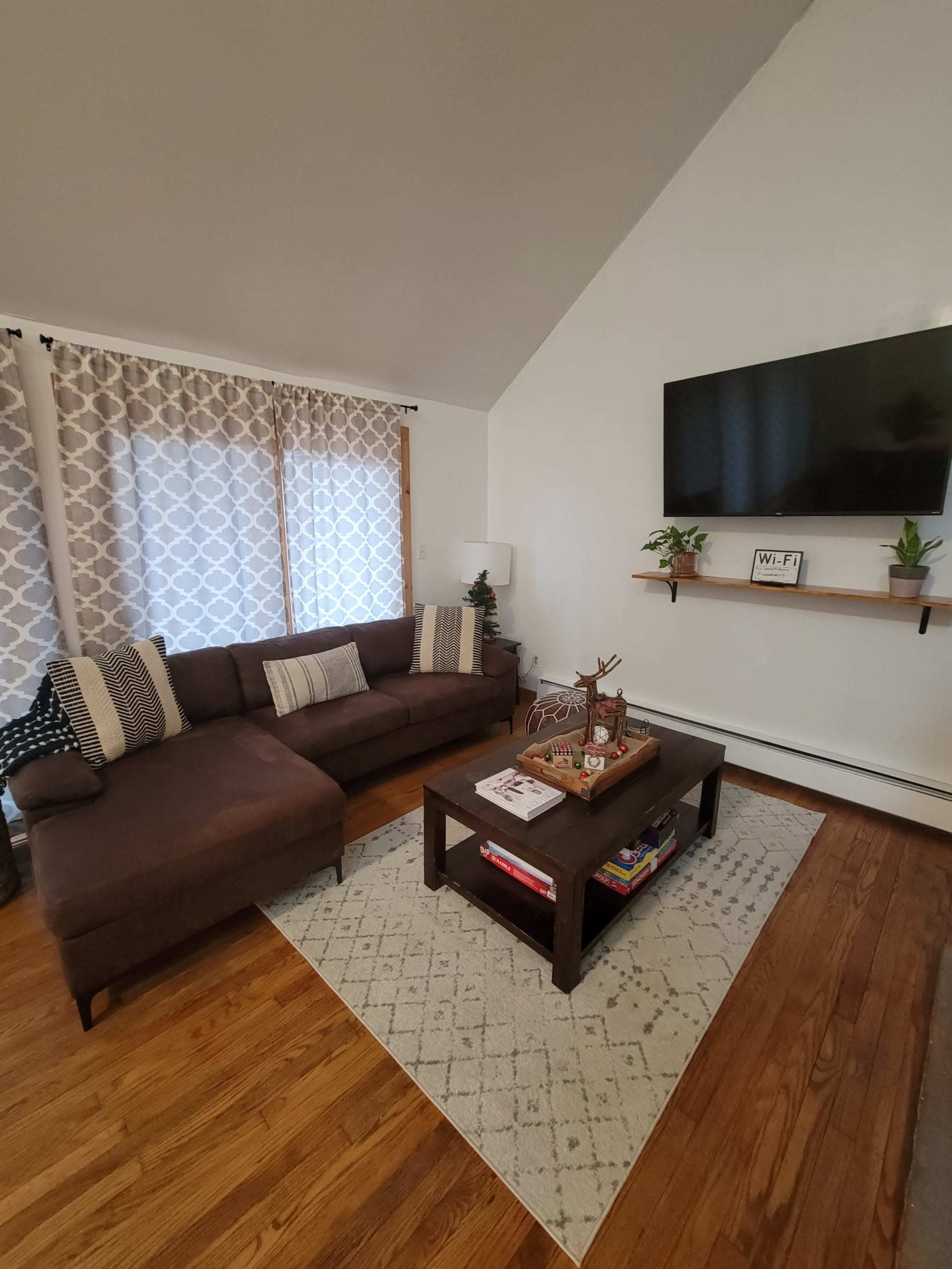 A living room with a brown sectional sofa, a coffee table on a rug, and a mounted television above a shelf with potted plants.