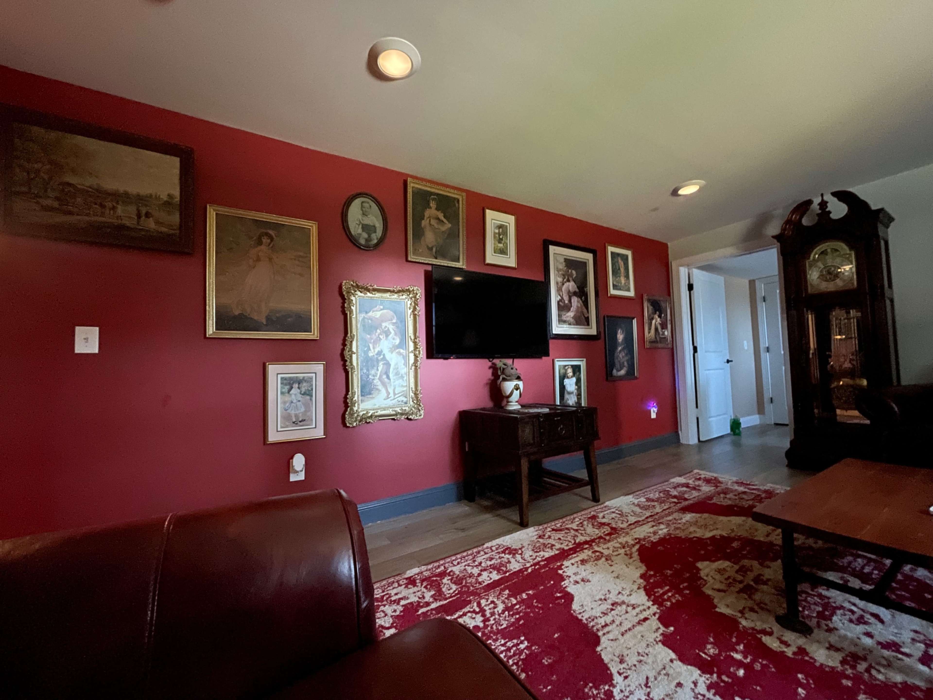 The image shows a living room with red walls adorned with various framed artworks and a television mounted on the wall, alongside a wooden cabinet and a patterned rug.