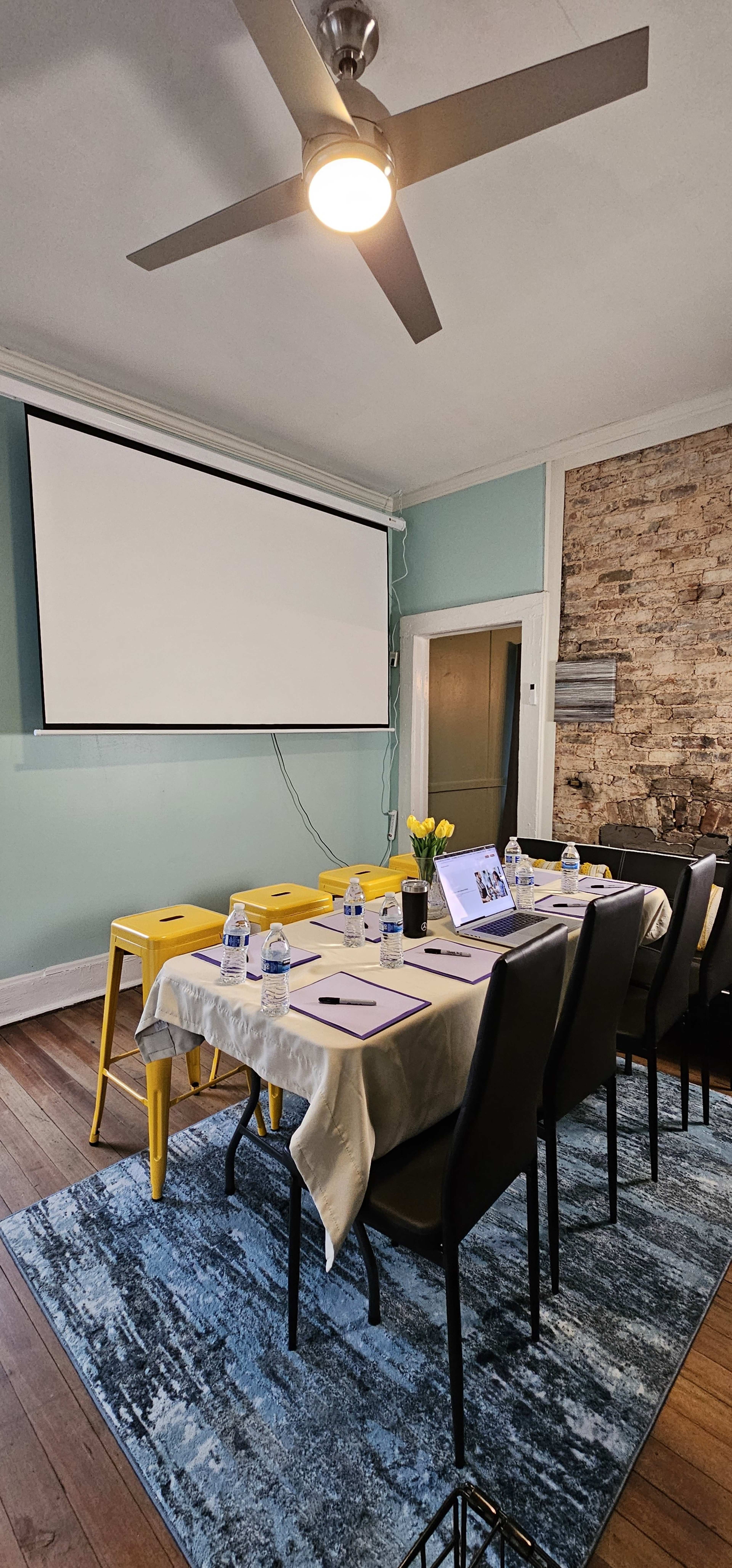 A meeting room is set up with a long table covered in a tablecloth, chairs around it, a projector screen at the front, and water bottles placed on the table.