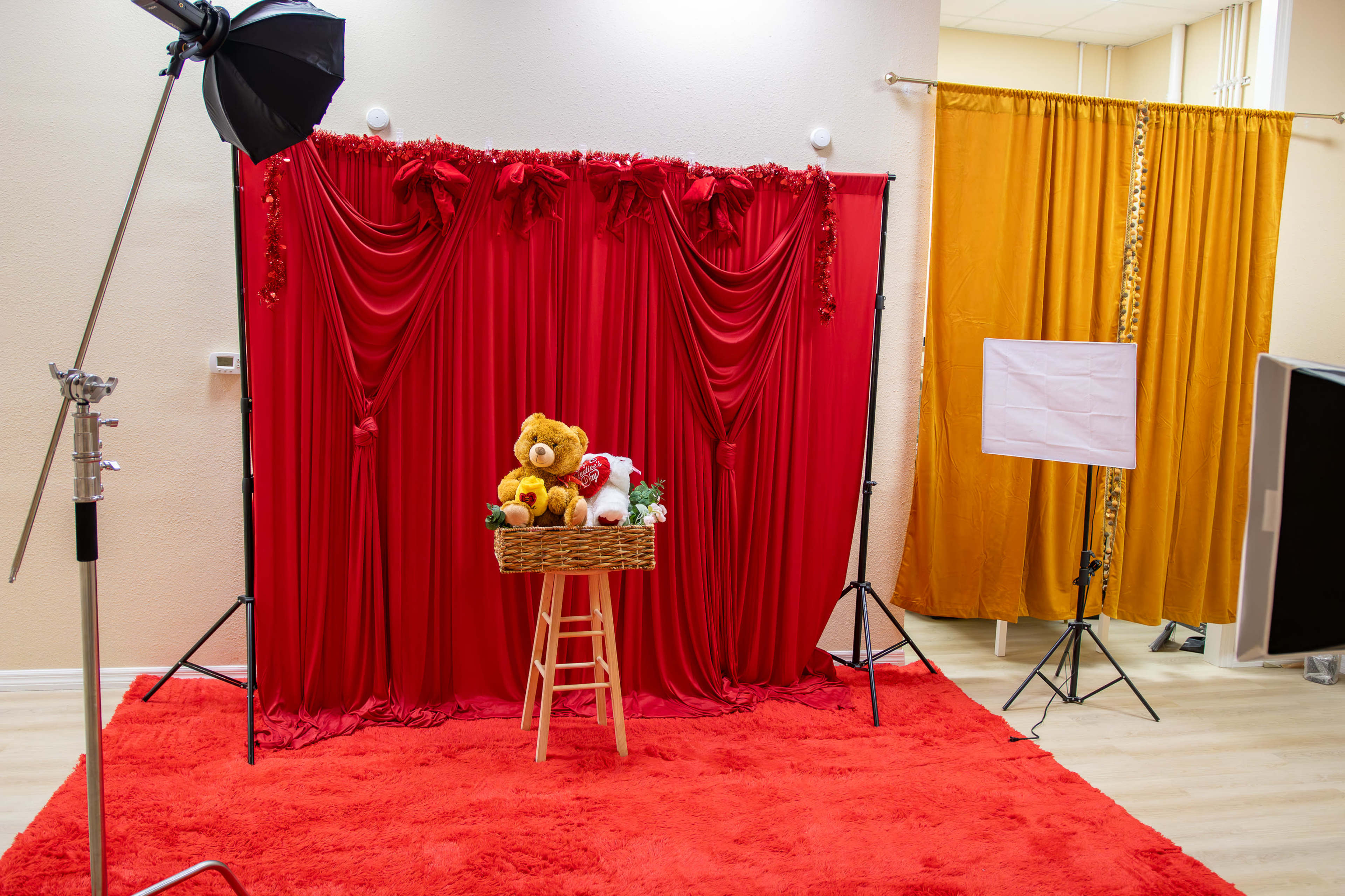 The image shows a photography studio setup with a red fabric backdrop, a basket of stuffed animals on a wooden stool in the center, and a yellow curtain on the side.