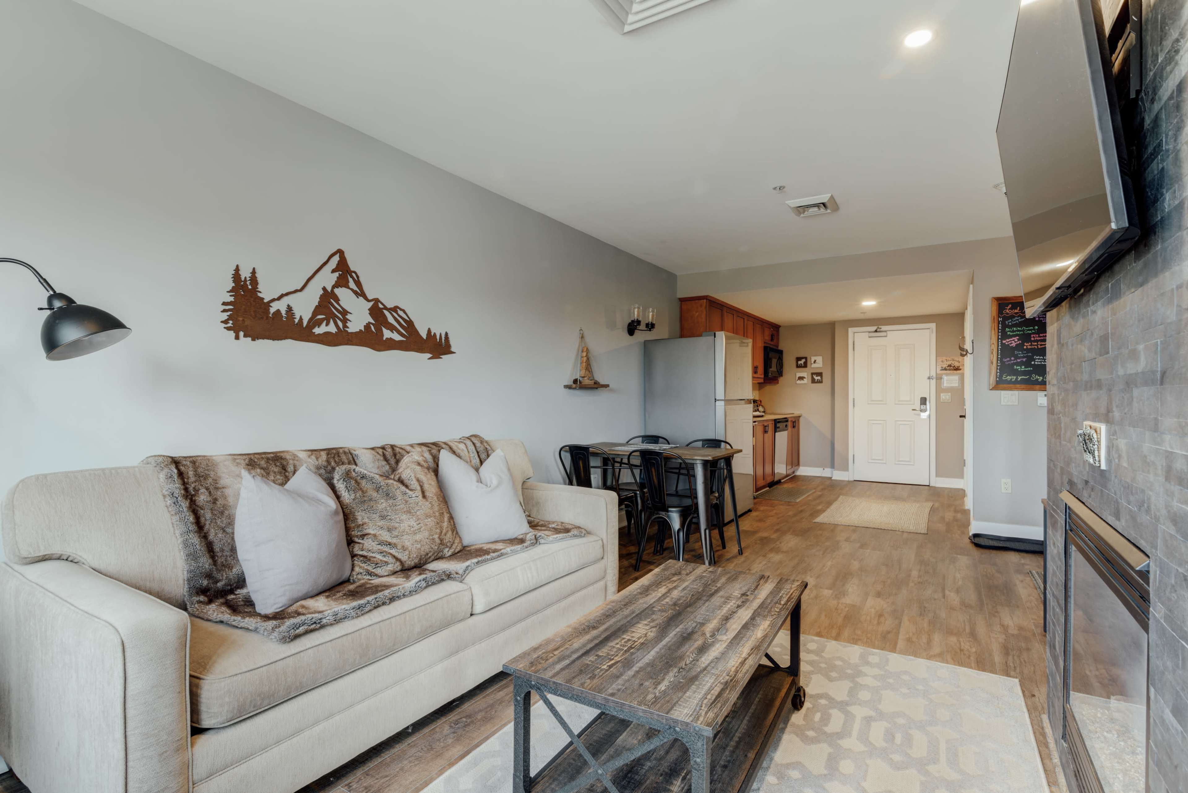 A modern living room with a beige sofa, a wooden coffee table, and a kitchenette in the background.
