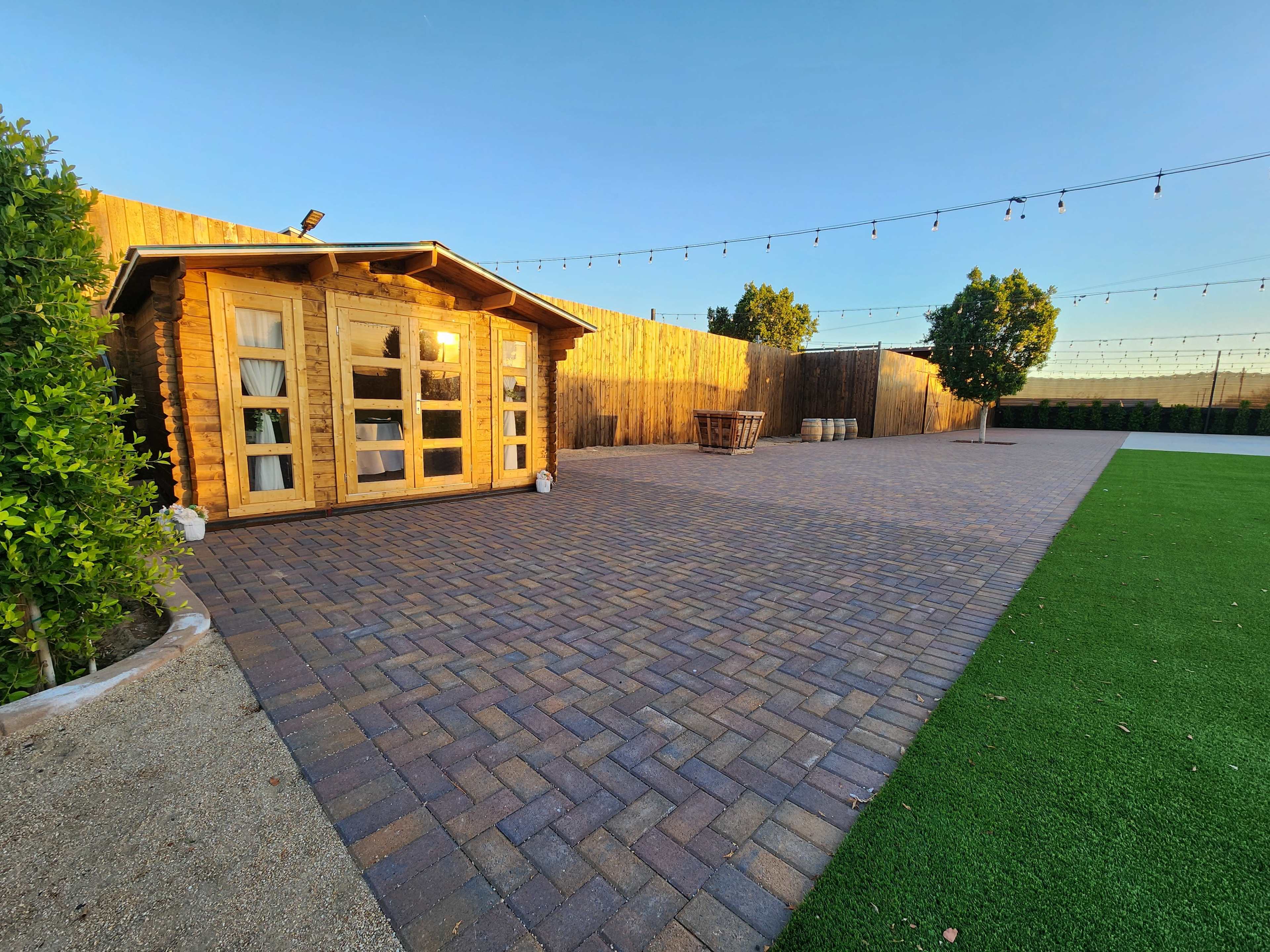 A wooden cabin next to a paved patio area surrounded by low hedges and illuminated by string lights.