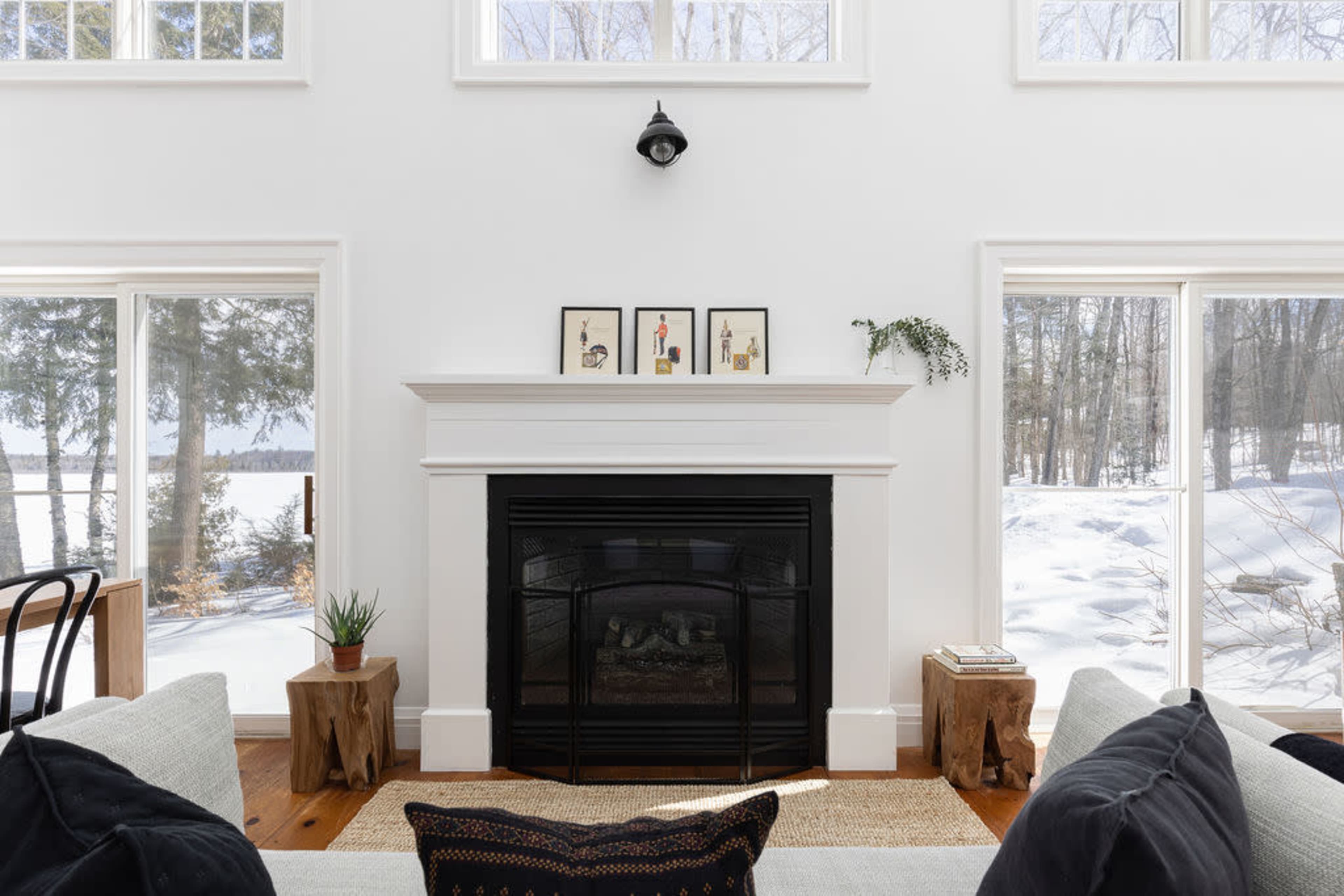 The cozy living room features a black fireplace surrounded by white walls, with large windows showing a snowy landscape outside.