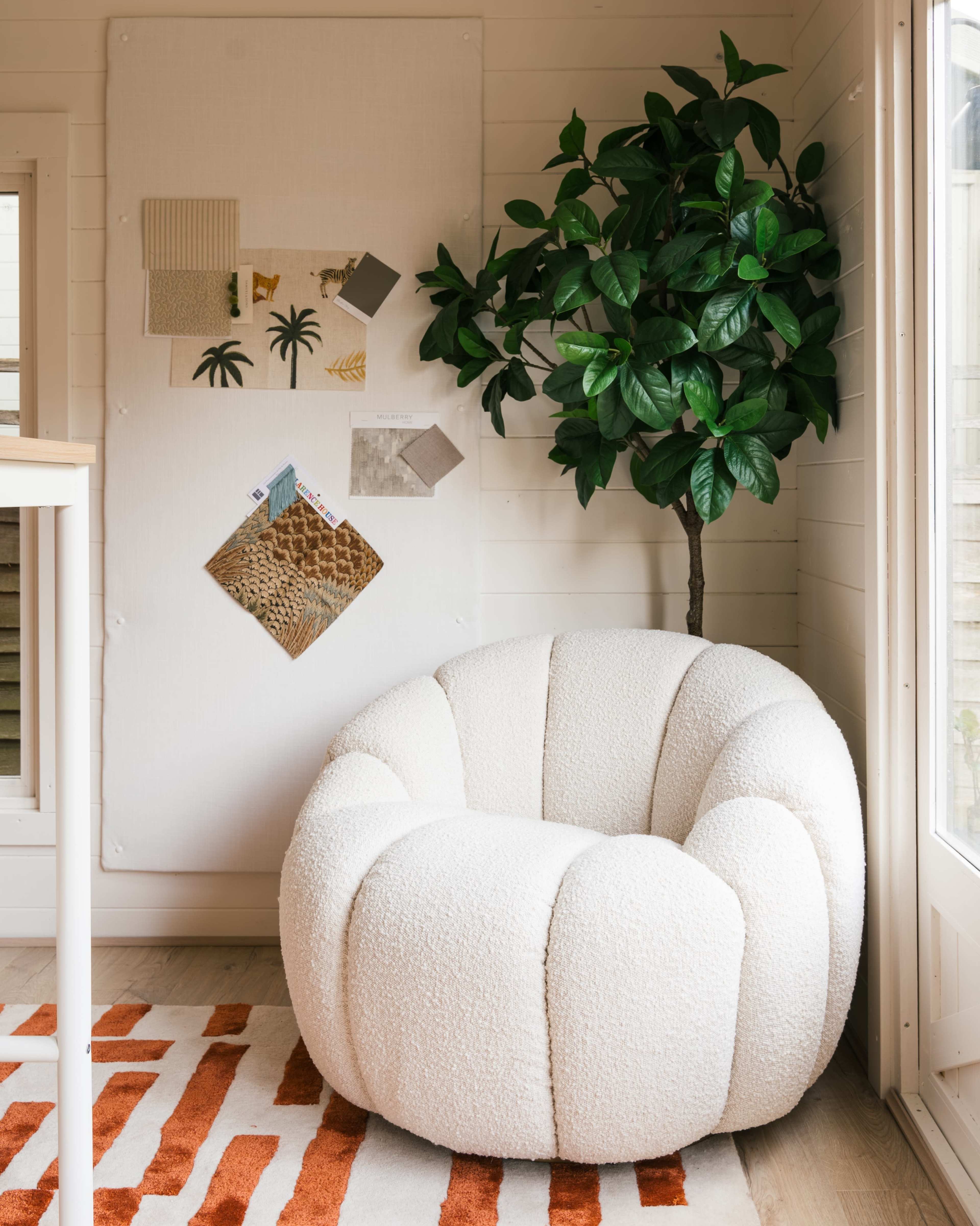 A rounded, textured white chair placed beside a green potted plant in a well-lit room with a collage board on the wall.