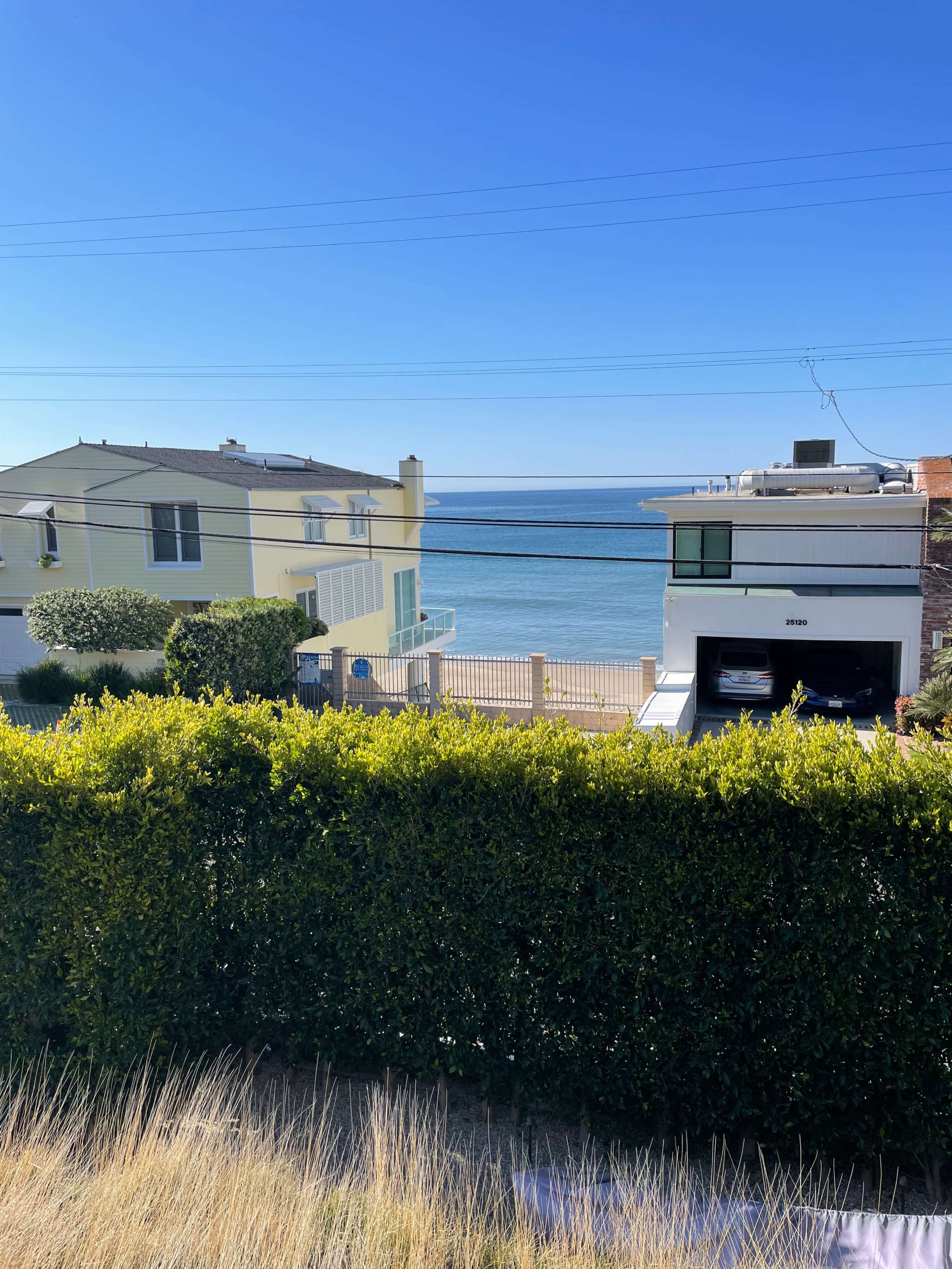 A view of the ocean framed by two houses and a hedge in the foreground.