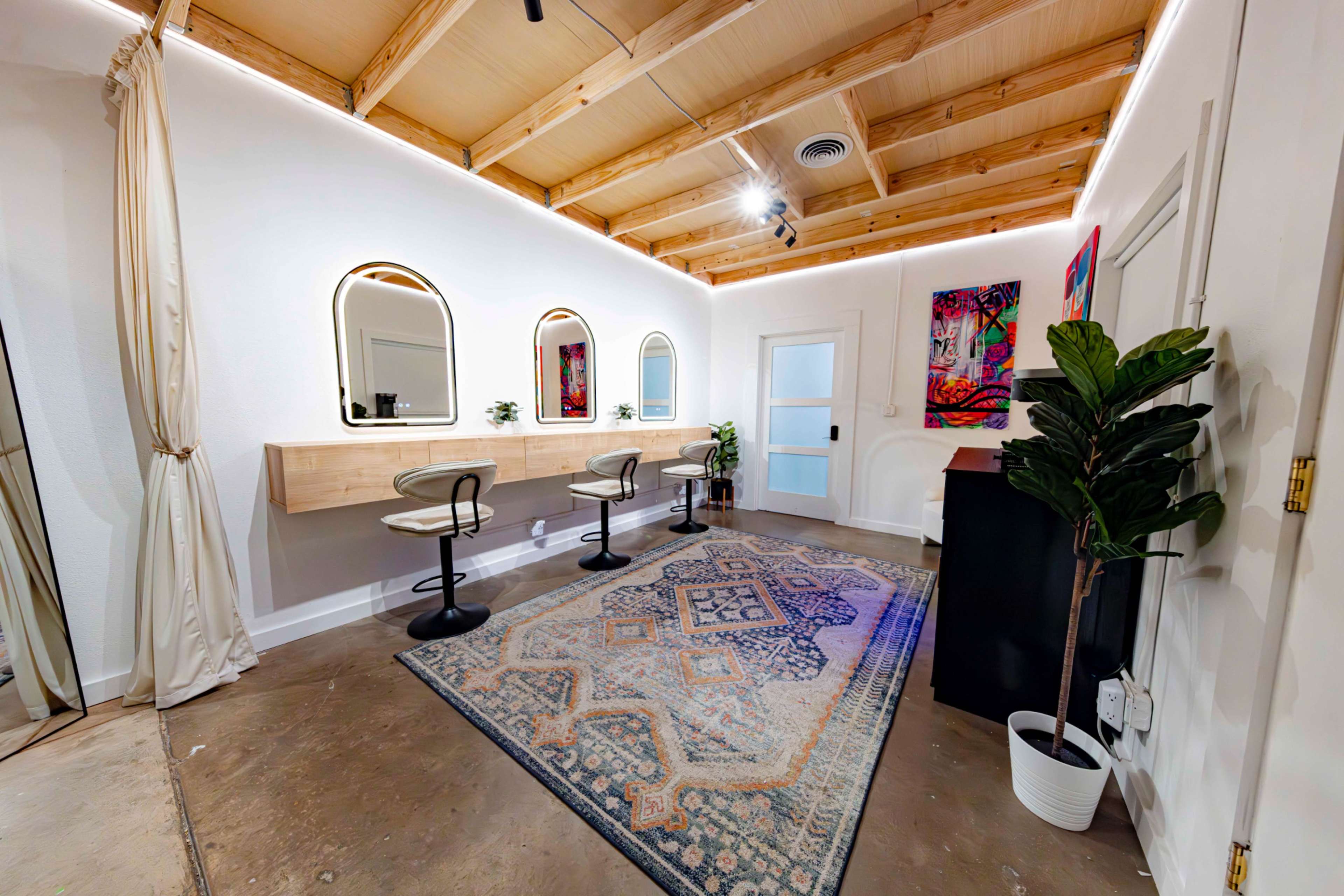 The image shows a modern beauty studio featuring three circular mirrors above wooden vanities, a decorative rug, and a potted plant, all set under a wooden ceiling.