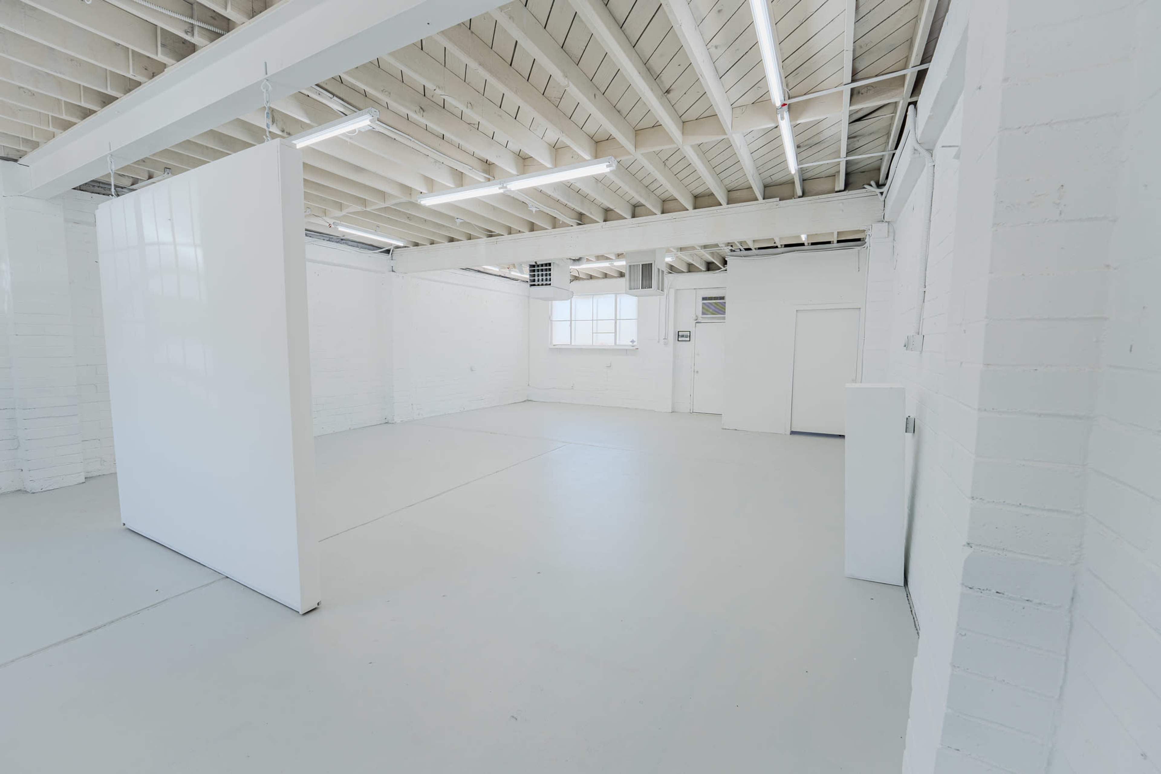An empty, well-lit room with white walls and a concrete floor, featuring exposed beams and fluorescent lighting.