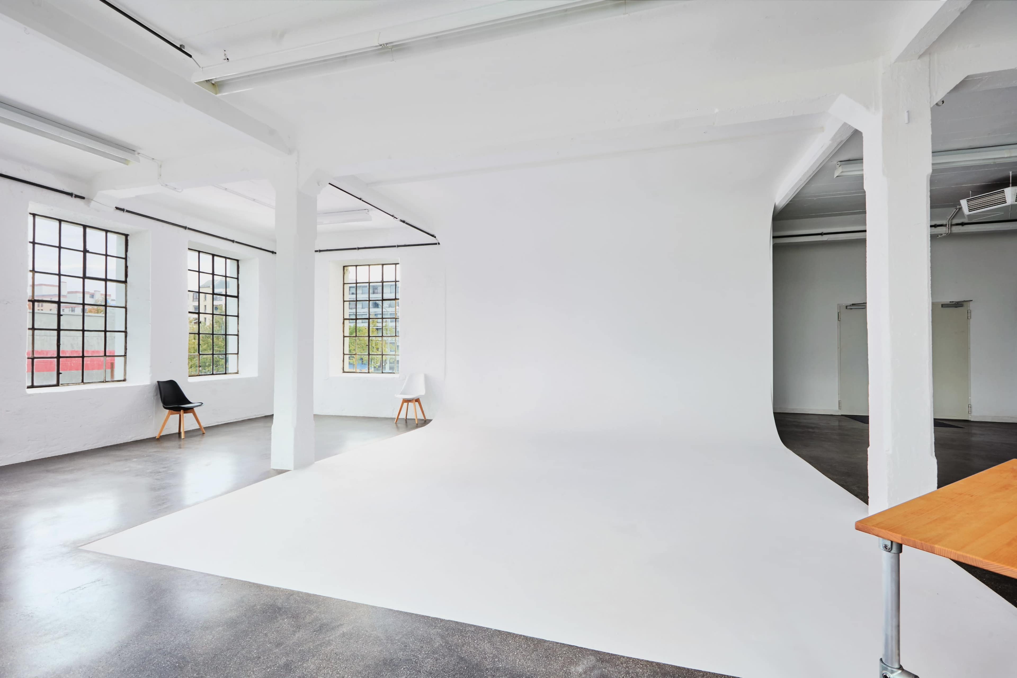 The image shows a large, empty photography studio with a white background and large windows letting in natural light.