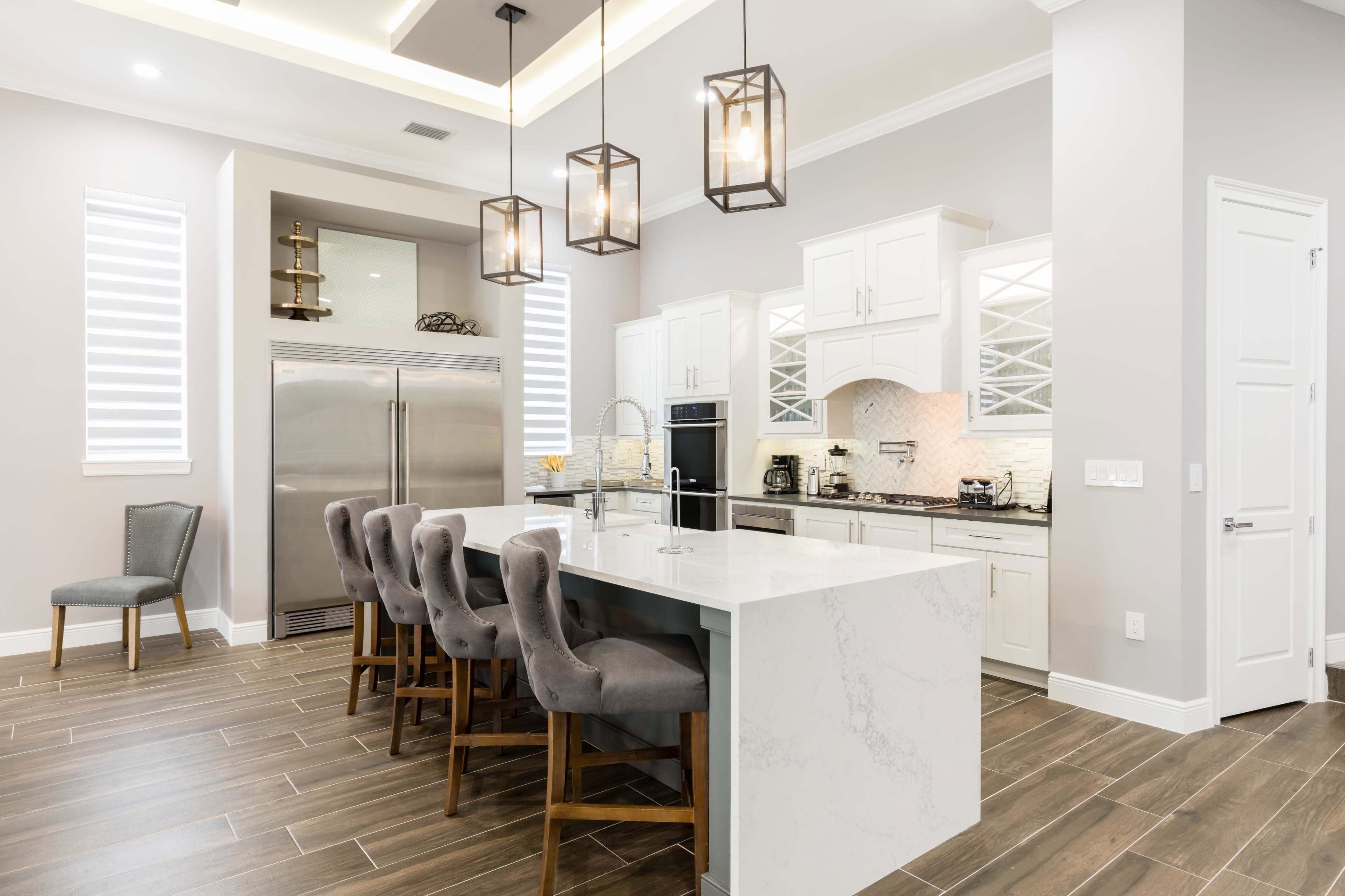 The image shows a modern kitchen with a central island, six bar stools, stainless steel appliances, and white cabinetry.